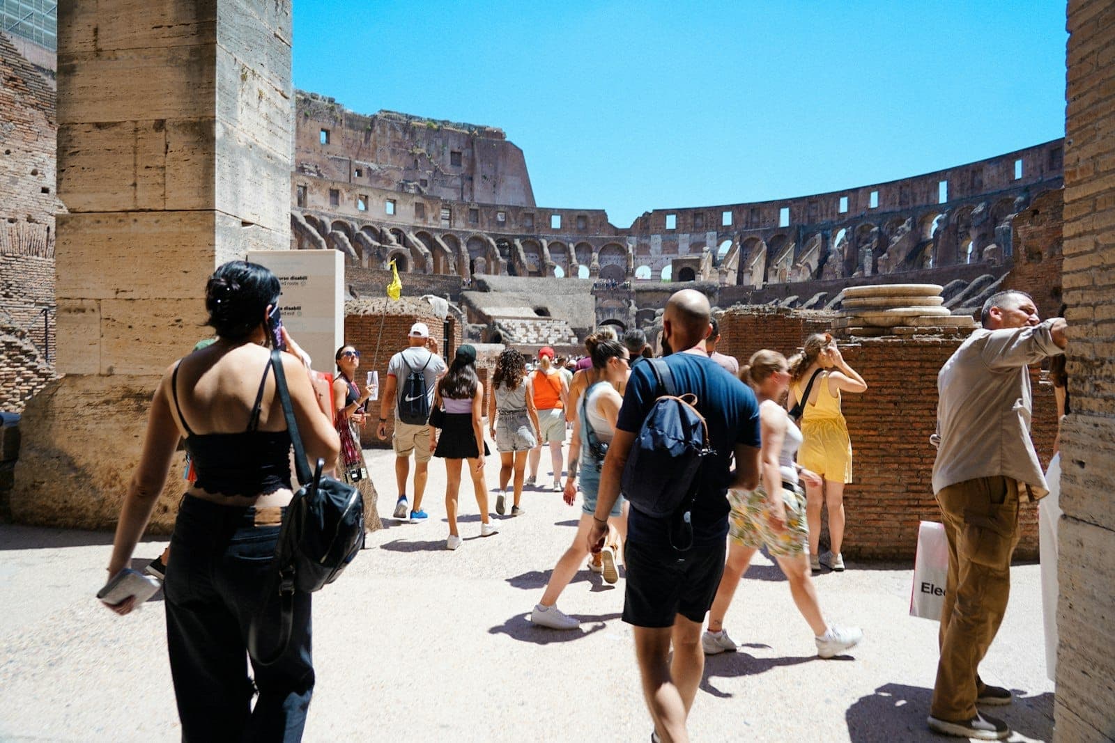 Wisatawan di dalam Colosseum yang diterangi sinar matahari mengenakan pakaian musim panas, dengan langit biru cerah dan dinding batu kuno yang terlihat.
