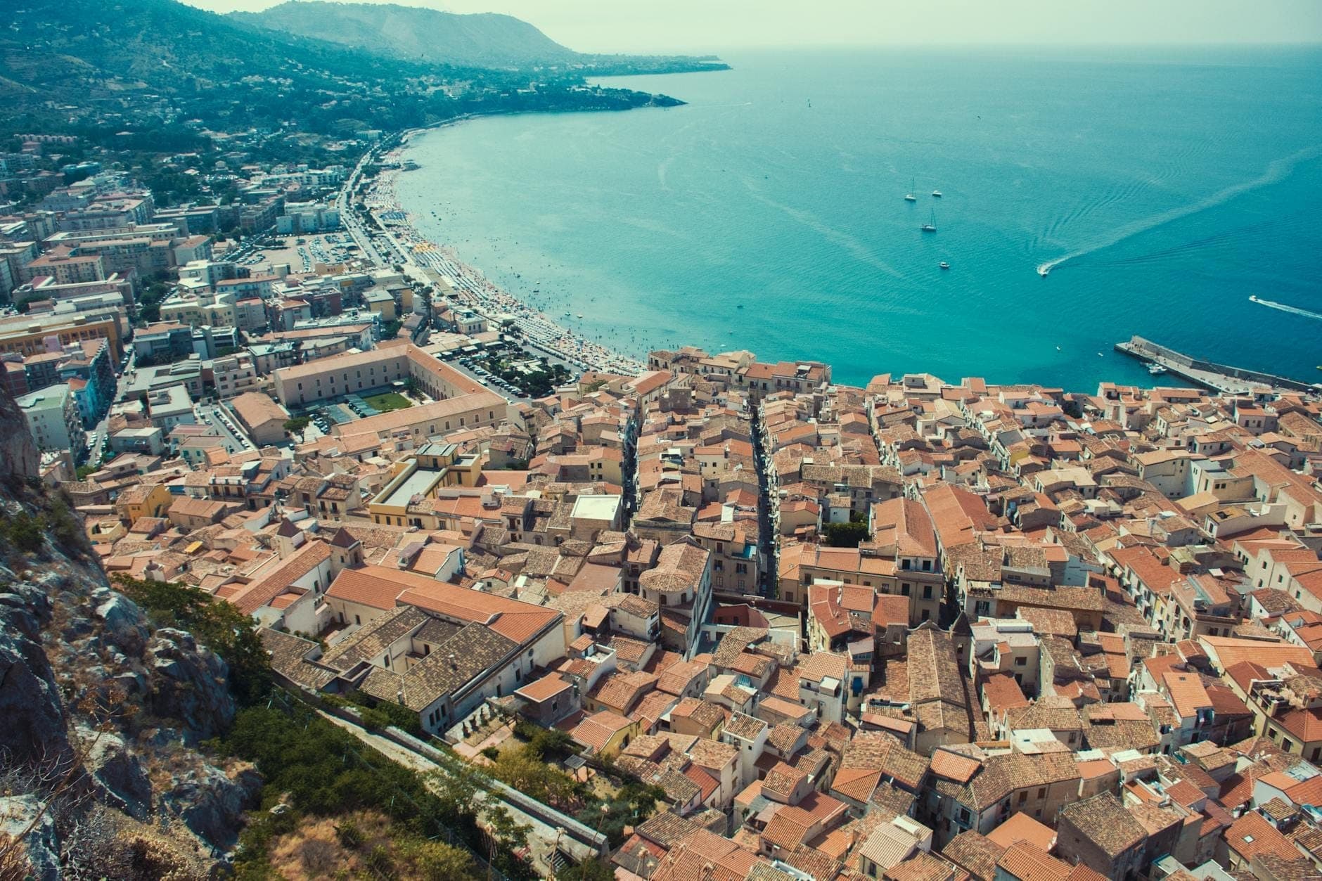Vista aerea della costa della Sicilia e del centro storico con tetti in terracotta, mare blu cristallino e spiaggia di sabbia curvilinea, nel luminoso sole d'inizio autunno.