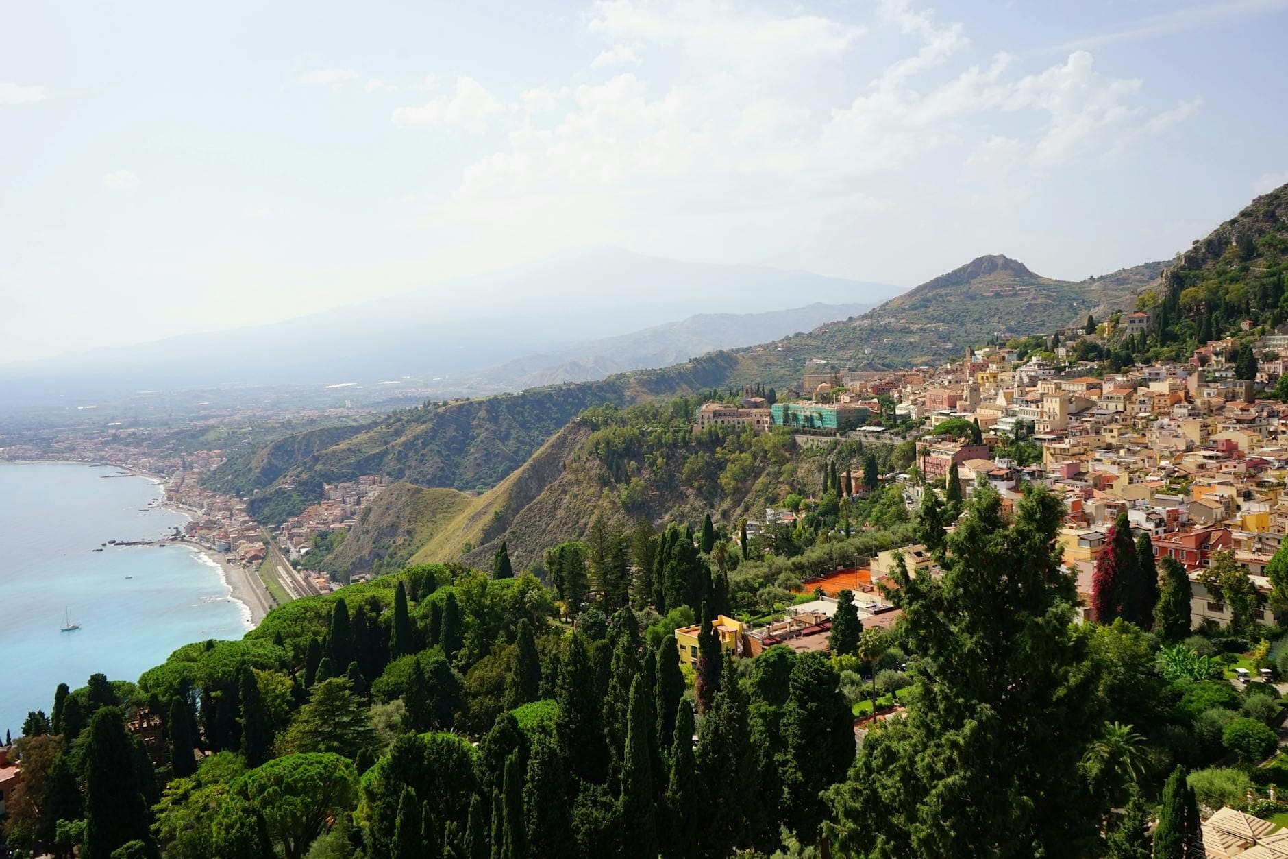 Vista costiera della Sicilia con mare turchese, rigogliosa vegetazione e un paese collinare sotto un cielo luminoso, con montagne lontane sullo sfondo.