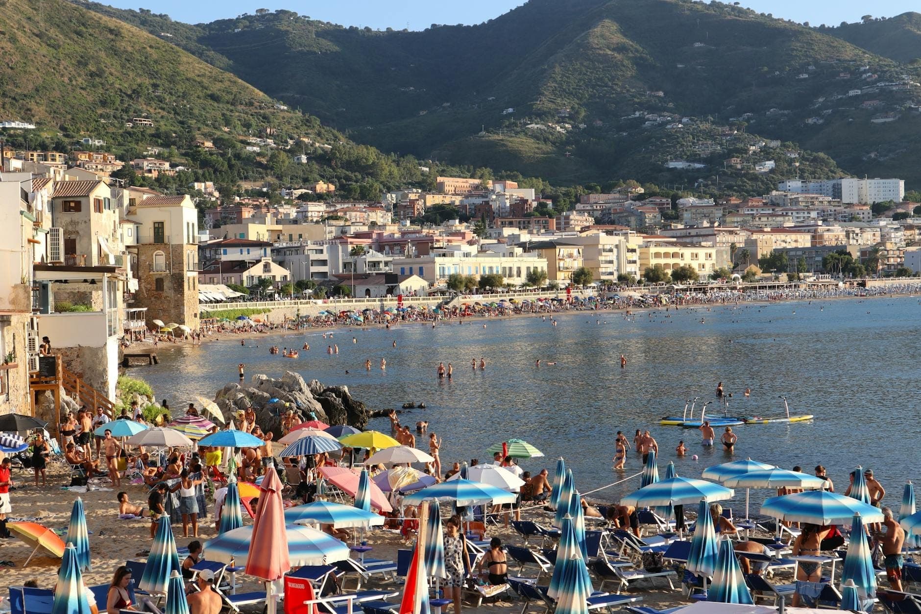 Affollata spiaggia siciliana in estate con ombrelloni, bagnanti, nuotatori in mare e colline con edifici di un paese sullo sfondo.