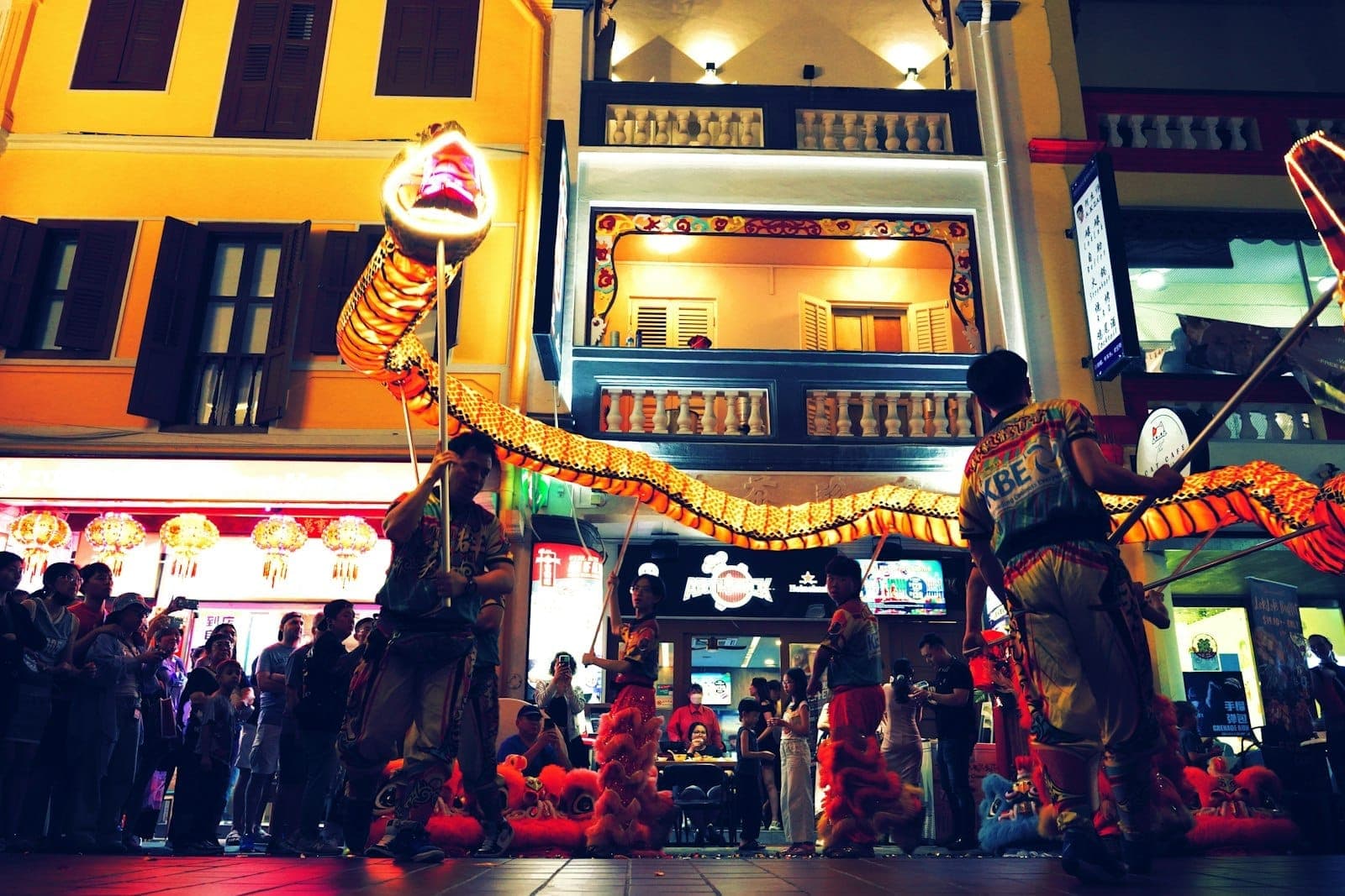 นักแสดงในชุดประเพณีถือมังกรเรืองแสงในขบวนพาเหรดยามค่ำคืนที่ไชนาทาวน์ของสิงคโปร์ โดยมีชอปเฮาส์หลากสีสันและโคมไฟเป็นฉากหลัง