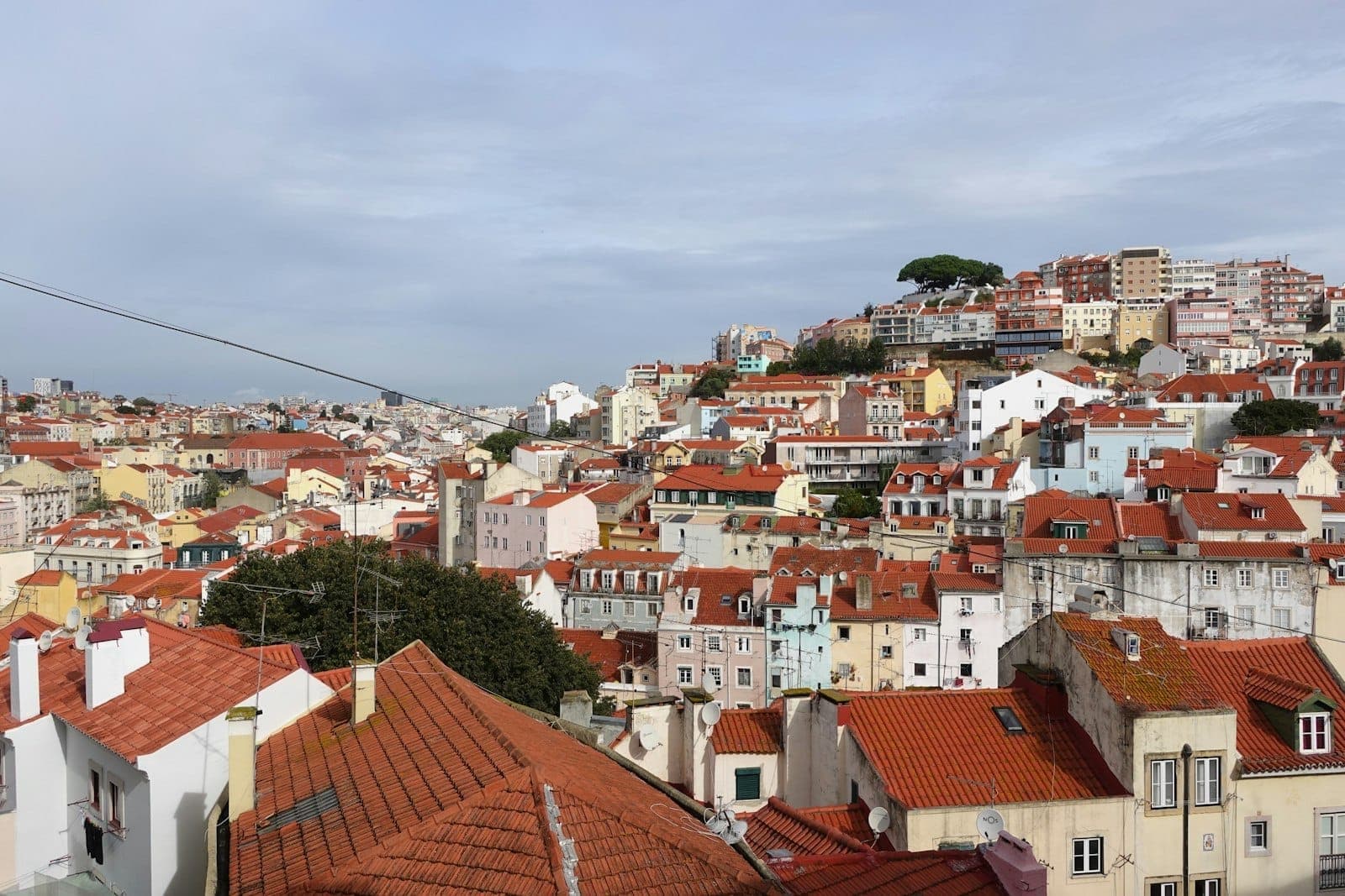 Vista ampla sobre as colinas de Alfama e Graça em Lisboa com casas coloridas, telhados de terracota e o horizonte ao fundo sob um céu nublado.
