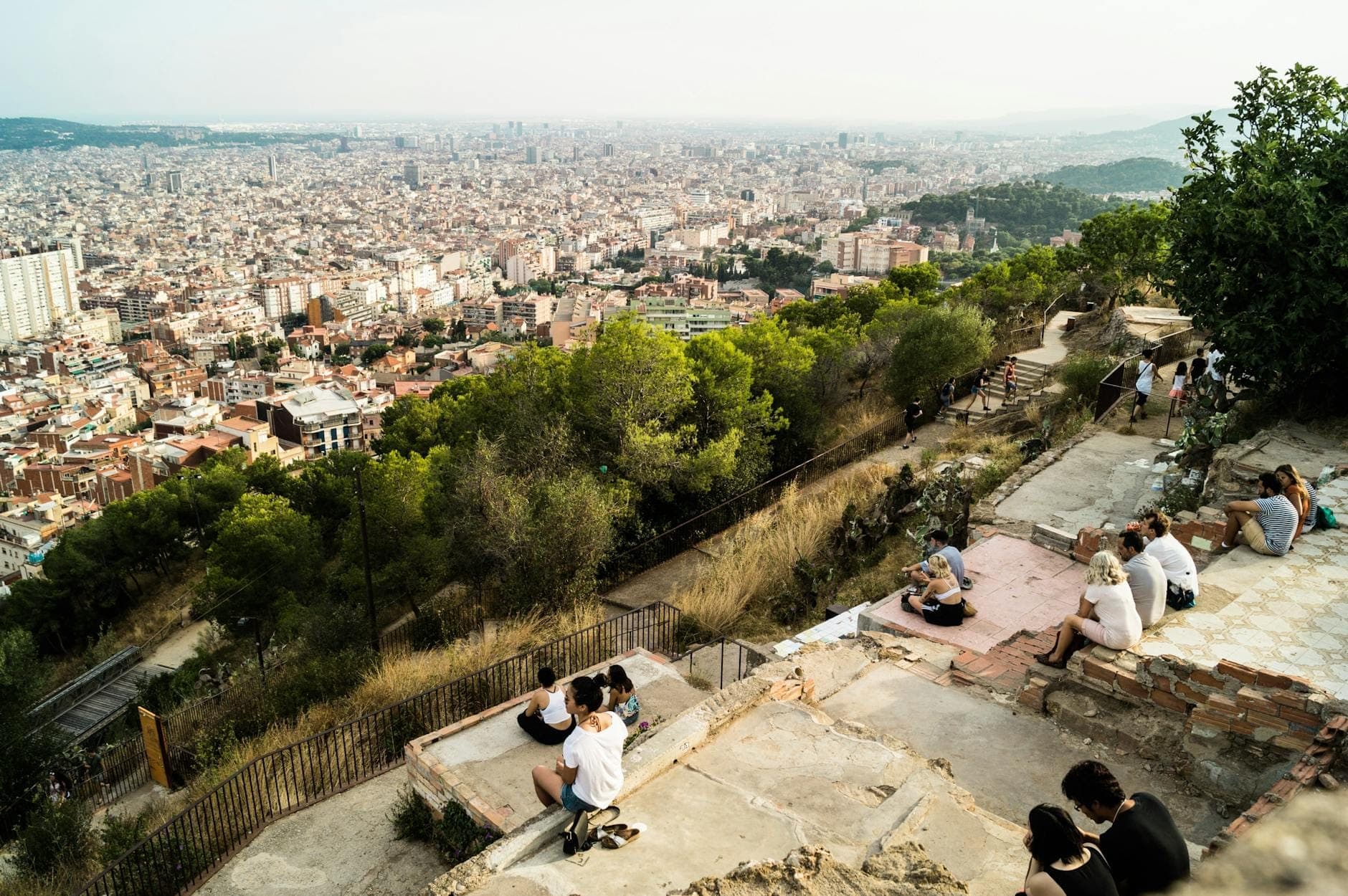 View from a hilltop overlooking Barcelona with people sitting on stone terraces, cityscape and Mediterranean Sea in the distance.