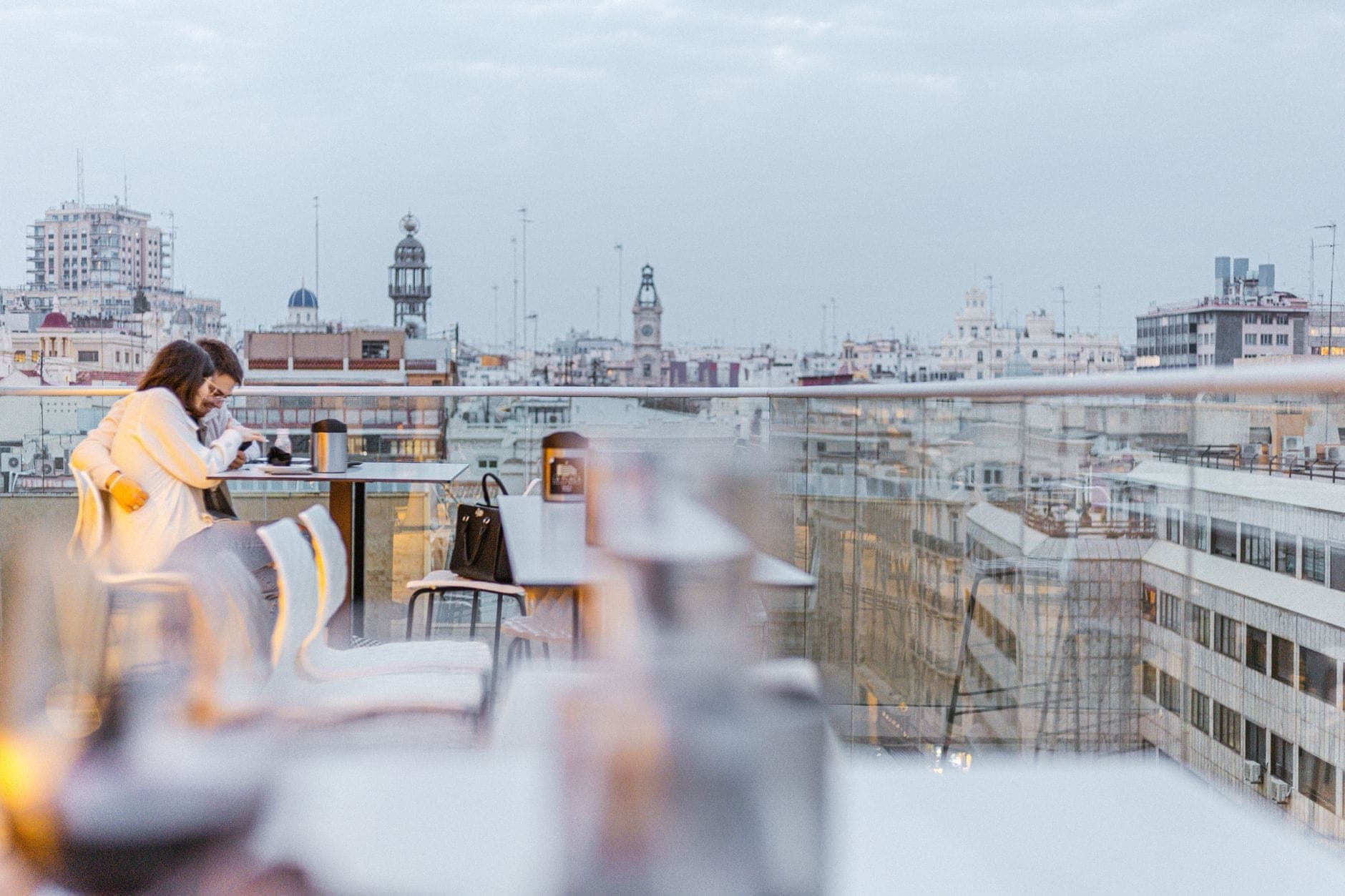 Moderne Dachterrassen-Bar in Madrid mit Glasbrüstungen, Tischen im Freien und der Skyline der Stadt im Hintergrund in der Abenddämmerung.