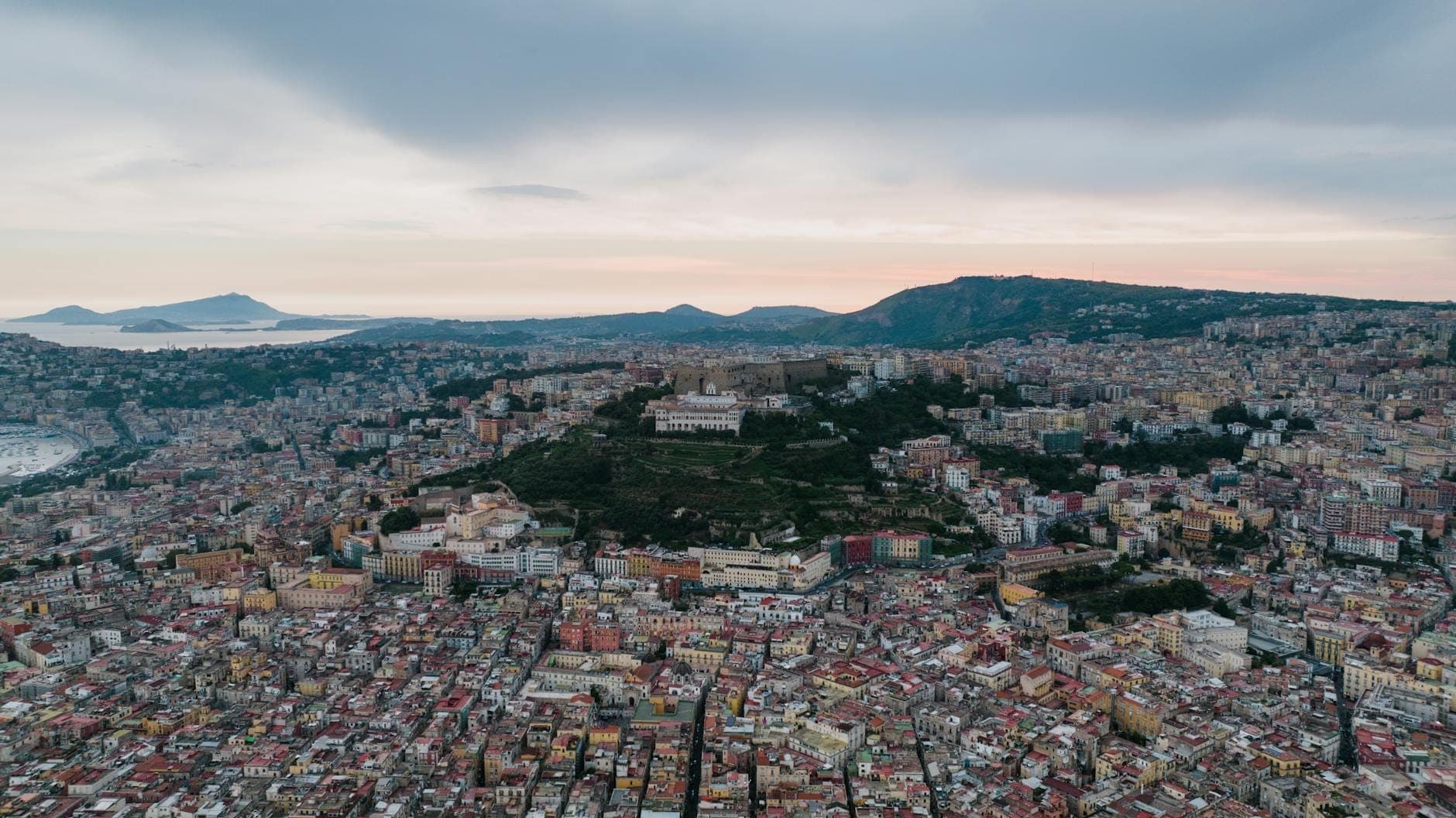 Vista aérea de Nápoles com o promontório de Posillipo avançando pela baía, colinas dramáticas, costa e o mar ao pôr do sol.