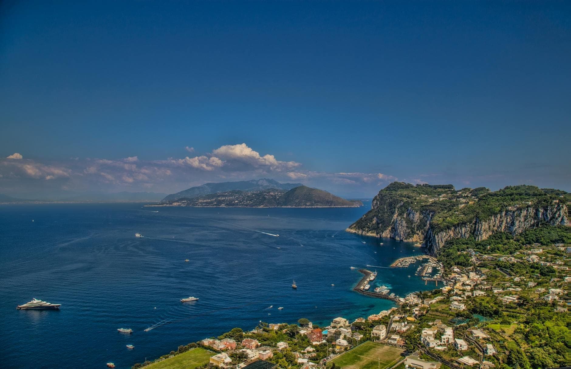 Vista dramática de um ponto alto sobre a baía, falésias e vila com barcos e formações terrestres distantes, típica da Baía de Nápoles e das ilhas próximas.