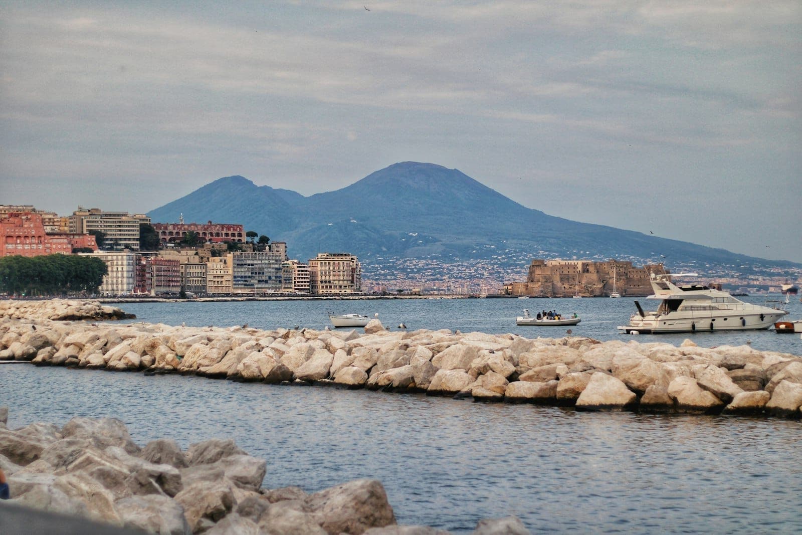 Vista do orla de Nápoles com edifícios, barcos, quebra-mar rochoso e o Monte Vesúvio ao fundo num dia nublado.