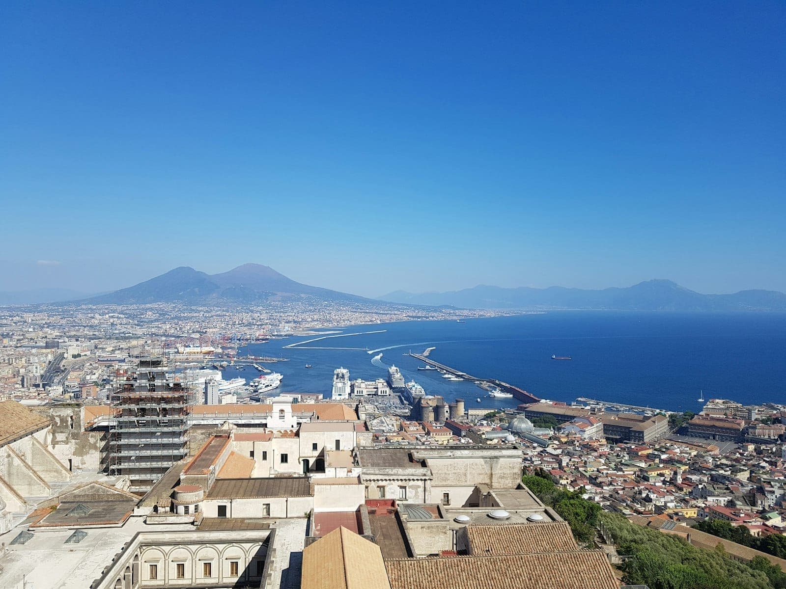 Vista panorâmica de um morro acima de Nápoles com o Monte Vesúvio, telhados da cidade, porto e um céu azul claro ao fundo.