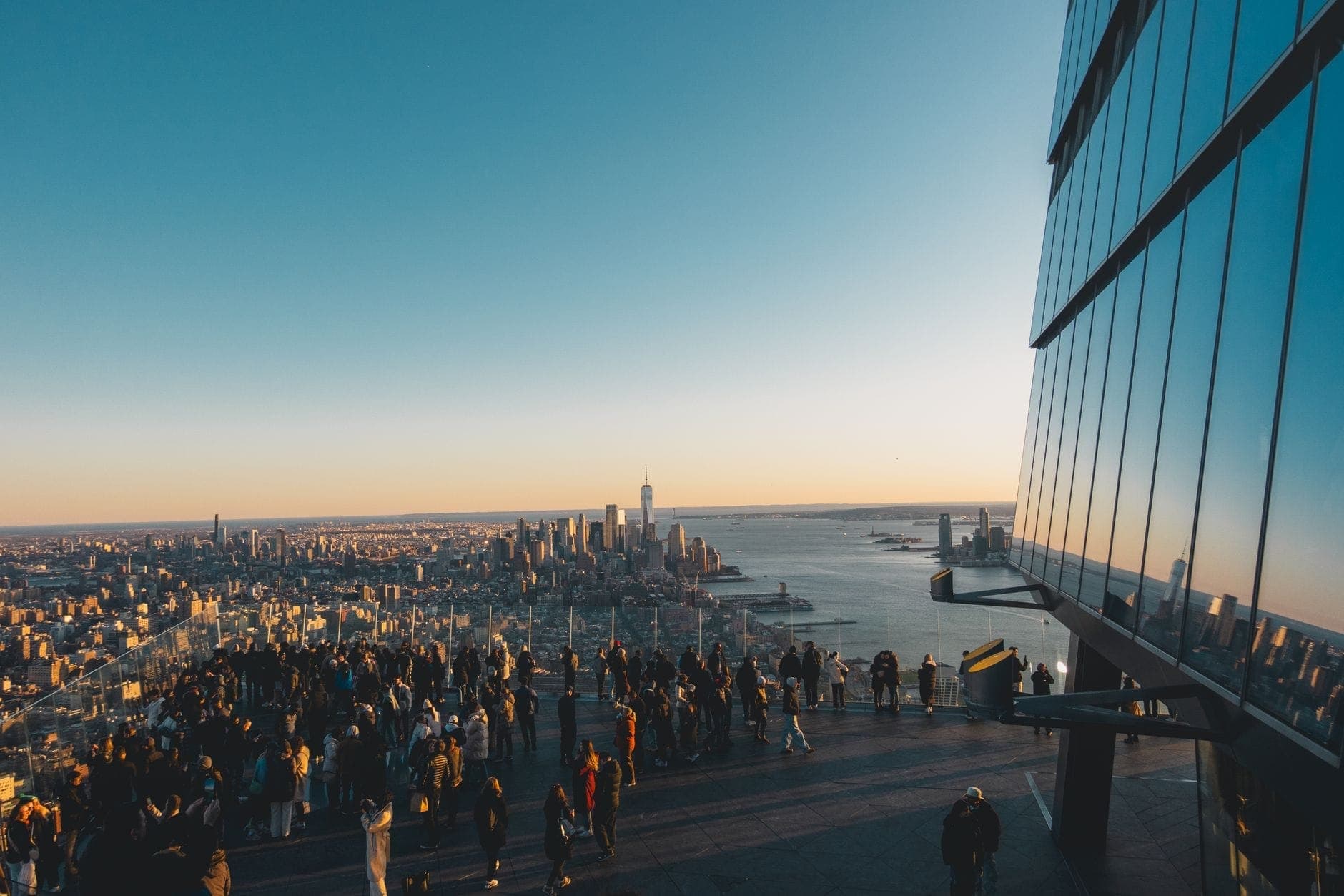 ผู้คนจำนวนมากเพลิดเพลินกับวิวแมนฮัตตันและ One World Trade Center จากดาดฟ้าเปิดโล่งช่วงพระอาทิตย์ตก เห็นเส้นขอบฟ้าเมืองและแม่น้ำฮัดสัน