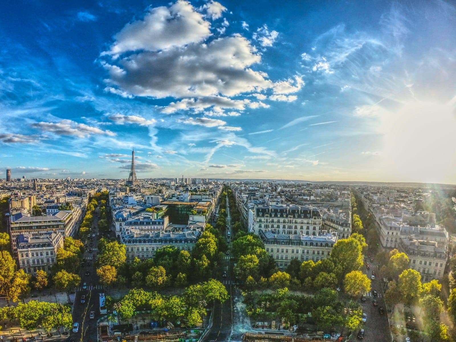 A dramatic bird’s-eye view of Paris beneath a bright blue sky with scattered clouds and the Eiffel Tower visible in the distance, sunlight casting a golden glow over the city’s avenues and rooftops.