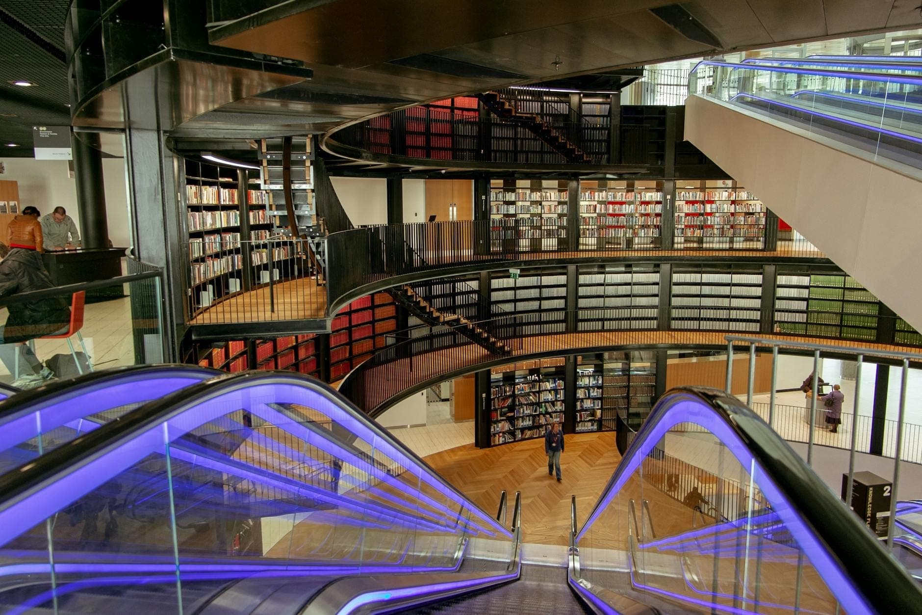 Modern interior view of a multi-level library with sweeping escalators, glass railings, and bookshelves lining the circular walls.