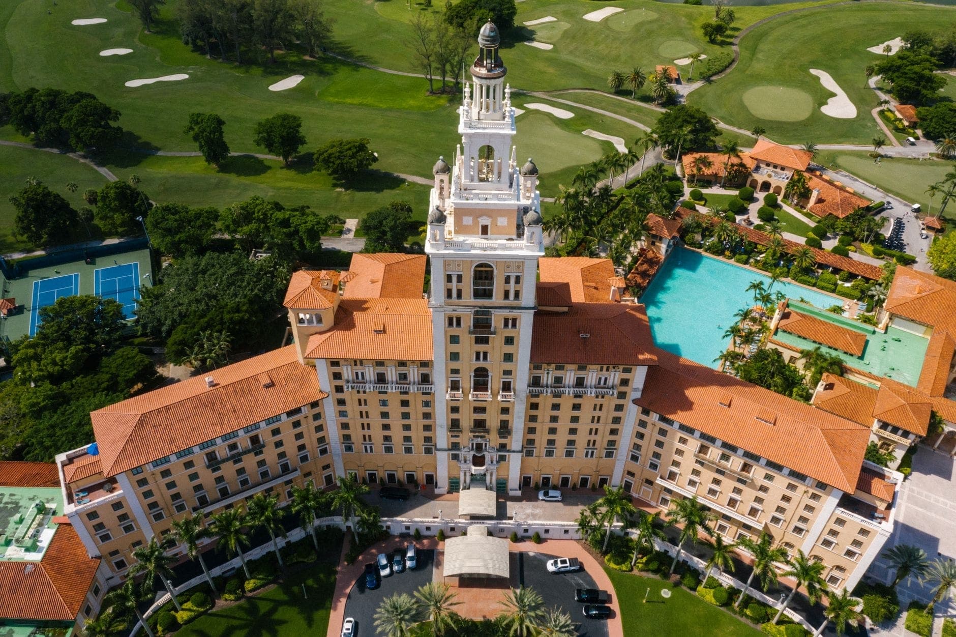 Aerial view of The Biltmore Hotel Coral Gables showing its iconic tower, red rooftops, lush gardens, and surrounding golf course on a sunny day.