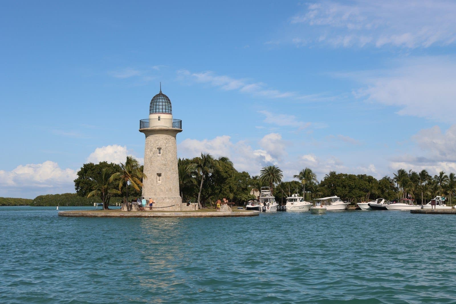 Stone lighthouse on Biscayne National Park's shore, surrounded by palm trees and boats on turquoise water under a sunny blue sky.
