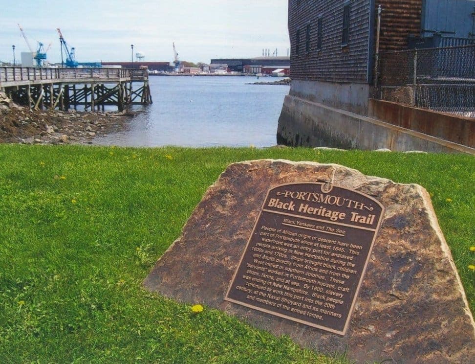 Une plaque en bronze du Portsmouth Black Heritage Trail fixée sur un rocher, avec pelouse, front de mer, jetée en bois et bâtiments industriels sous un ciel bleu.