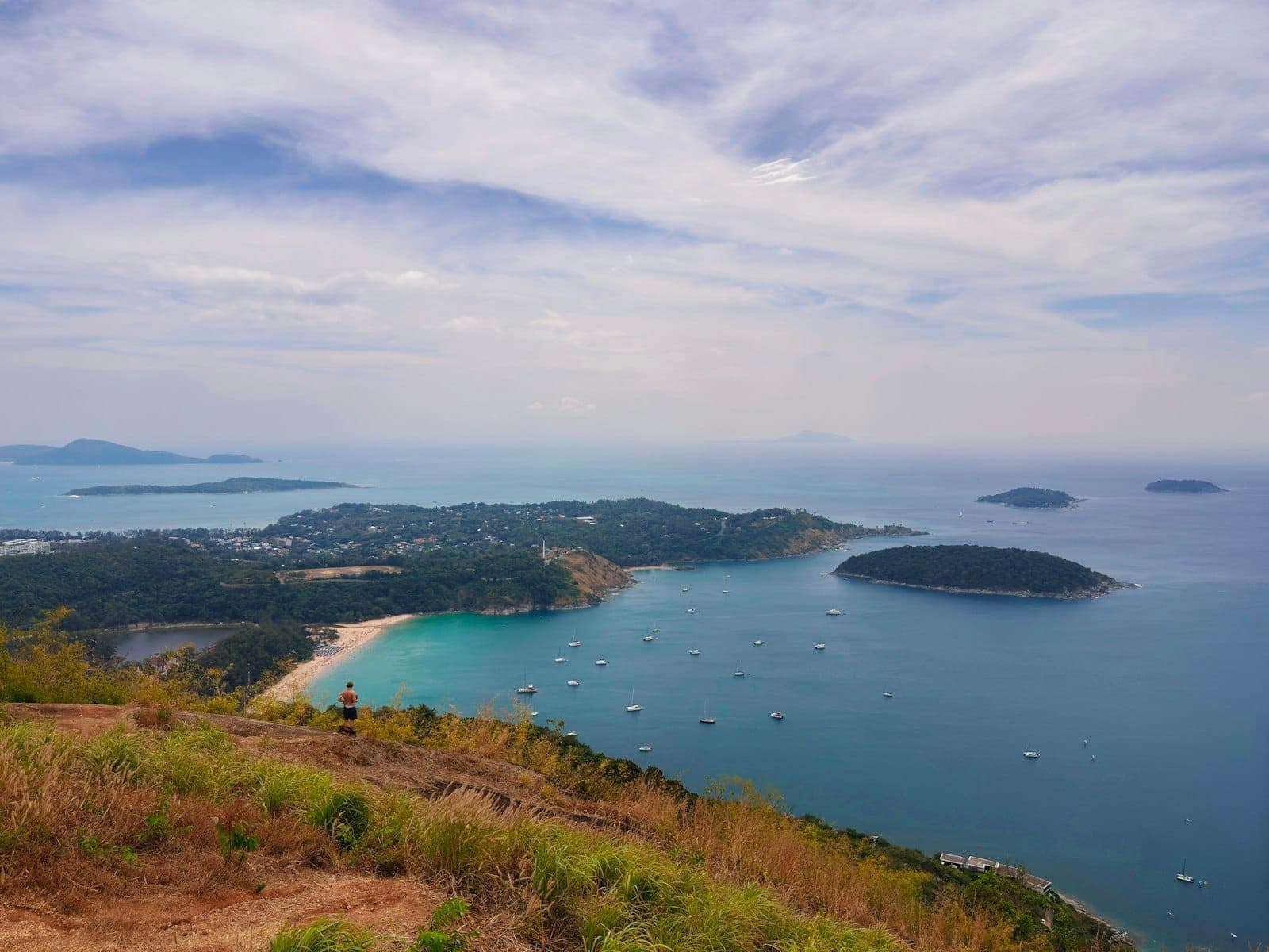 Wide panoramic view from Black Rock Viewpoint in Phuket, showing turquoise sea, green islands, Nai Harn Beach, and scattered boats under a cloudy sky.