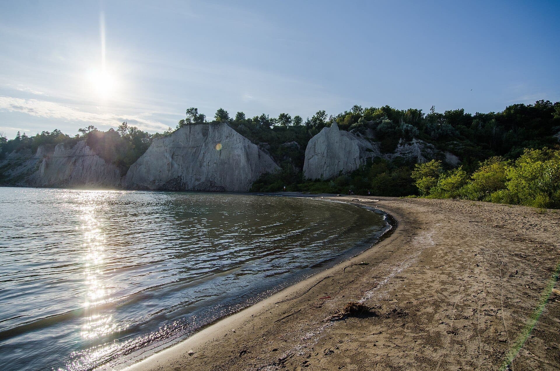 Una riva sabbiosa al Bluffer's Park and Beach con acque lacustri calme e le imponenti Scarborough Bluffs sotto un cielo limpido e soleggiato.