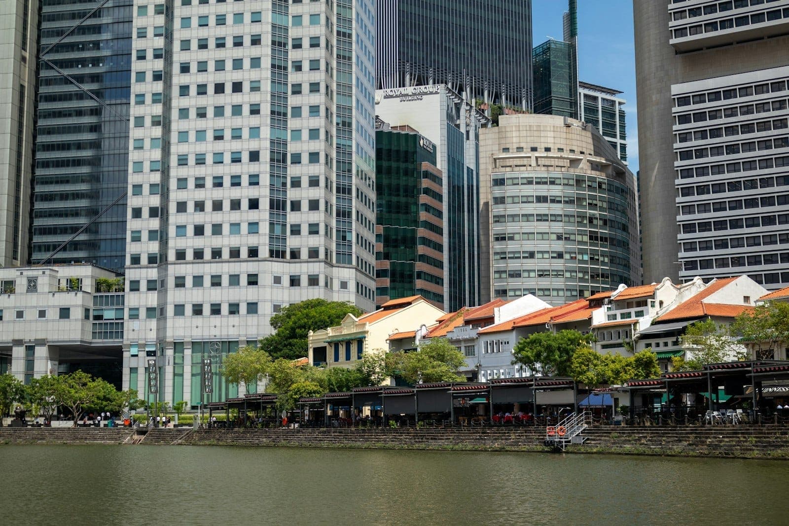 ตึกแถว Boat Quay ริมแม่น้ำสิงคโปร์ มีตึกระฟ้าสมัยใหม่เป็นฉากหลัง พร้อมร้านอาหารริมน้ำและต้นไม้เขียวขจีใต้ท้องฟ้าสดใส