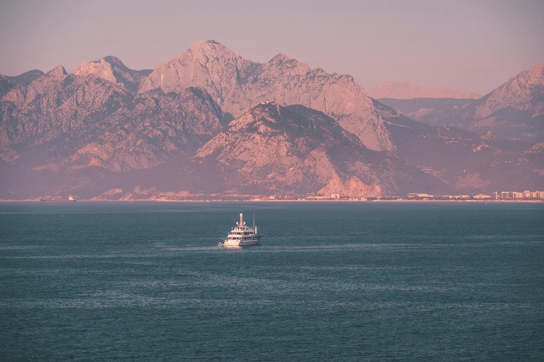 A boat cruising on the Mediterranean Sea with Antalya's dramatic mountains and city skyline in the background at sunset.