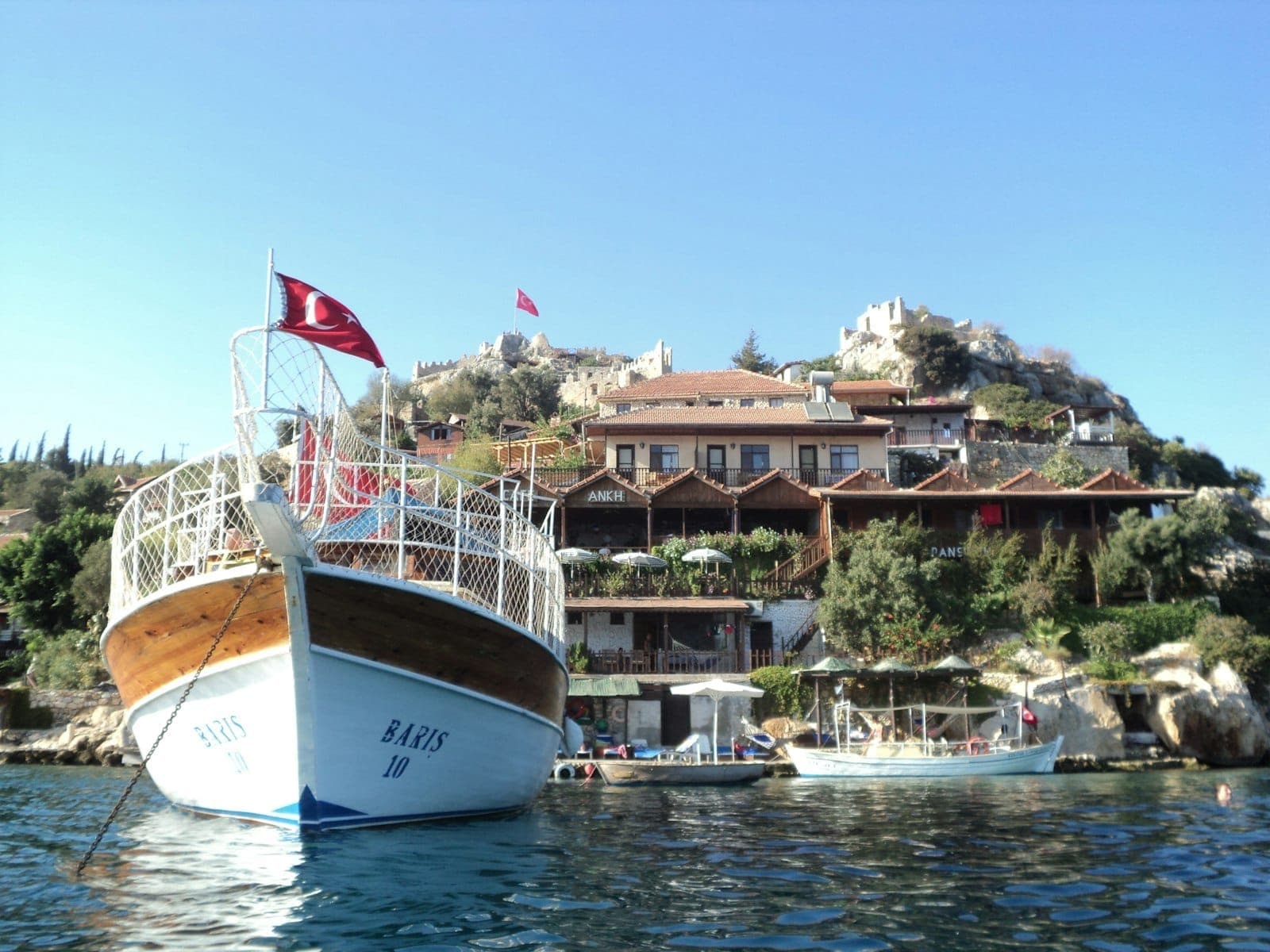 Ein hölzernes Touristenboot mit türkischer Flagge, das am Kaleiçi Marina liegt, mit Hangvillen und antiken Ruinen unter einem klaren blauen Himmel.