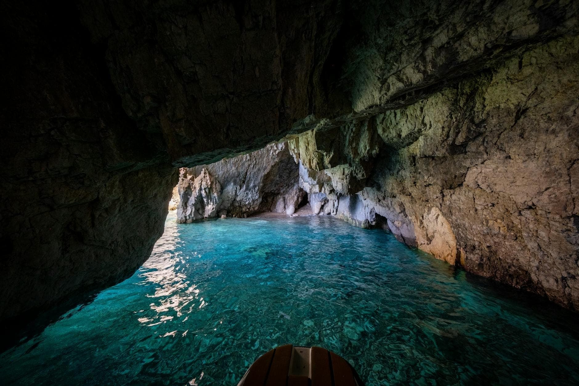 Vista dall'interno di una grotta marina con acqua turchese brillante e pareti di roccia naturale, verso l'ingresso e il mare illuminato dal sole.