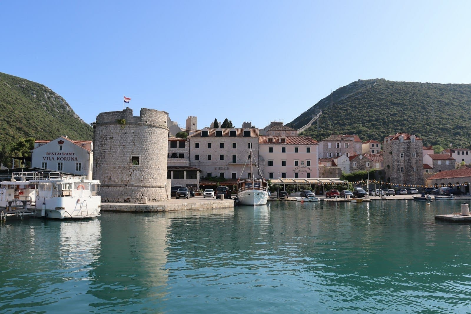 Piccolo porto con barche, torre in pietra e vecchi edifici sullo sfondo delle colline; probabilmente un villaggio su un'isola dalmata.