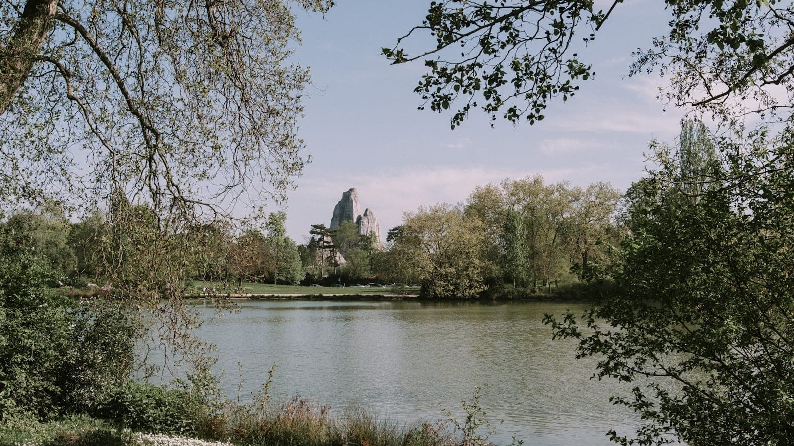 Tranquil lake surrounded by lush greenery and trees in the Bois de Vincennes, with a view of a castle-like structure in the distance under a bright sky.