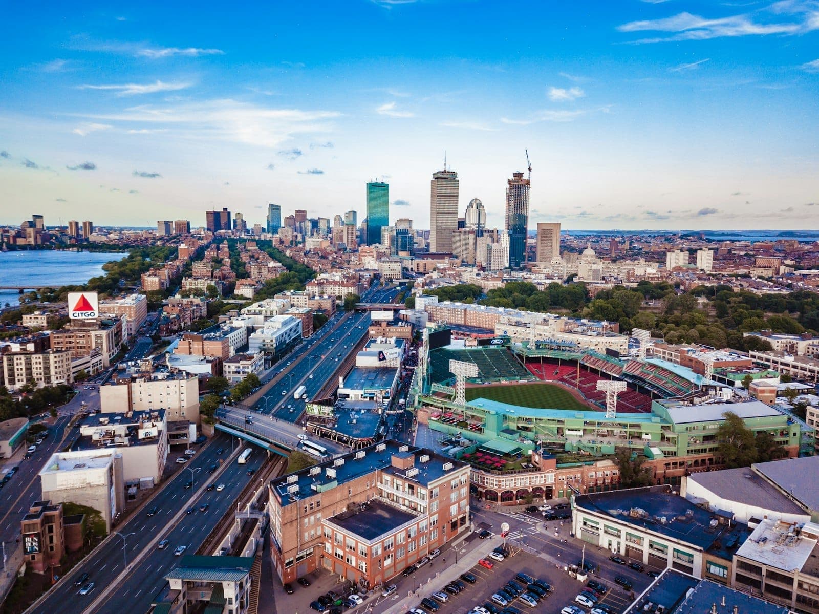 Aerial view of Boston skyline with Fenway Park in the foreground, highways, and the city’s iconic skyscrapers under a blue sky.