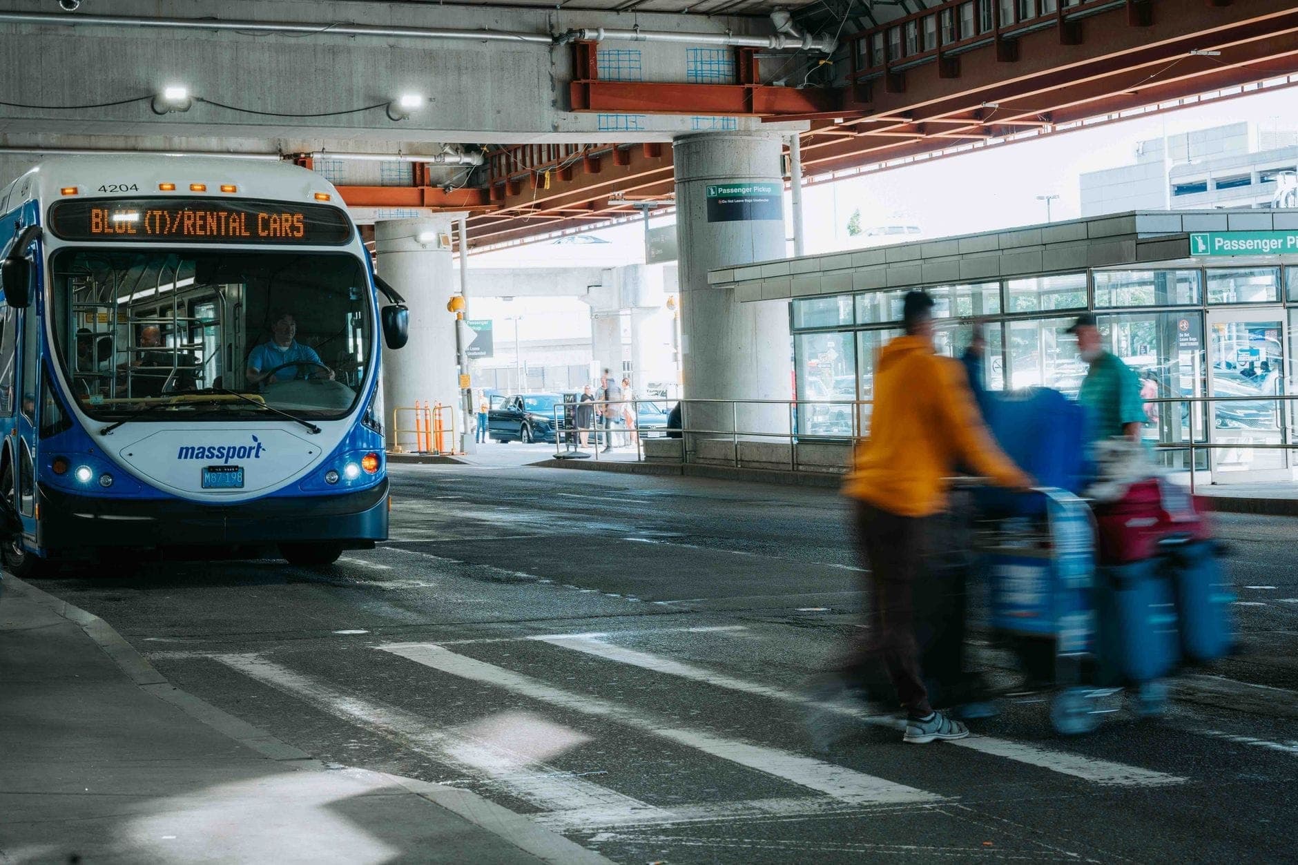 Un bus de navette Massport indiquant 'Rental Cars' dans la zone de transport terrestre de l'aéroport Boston Logan, avec des voyageurs et des bagages visibles à proximité.