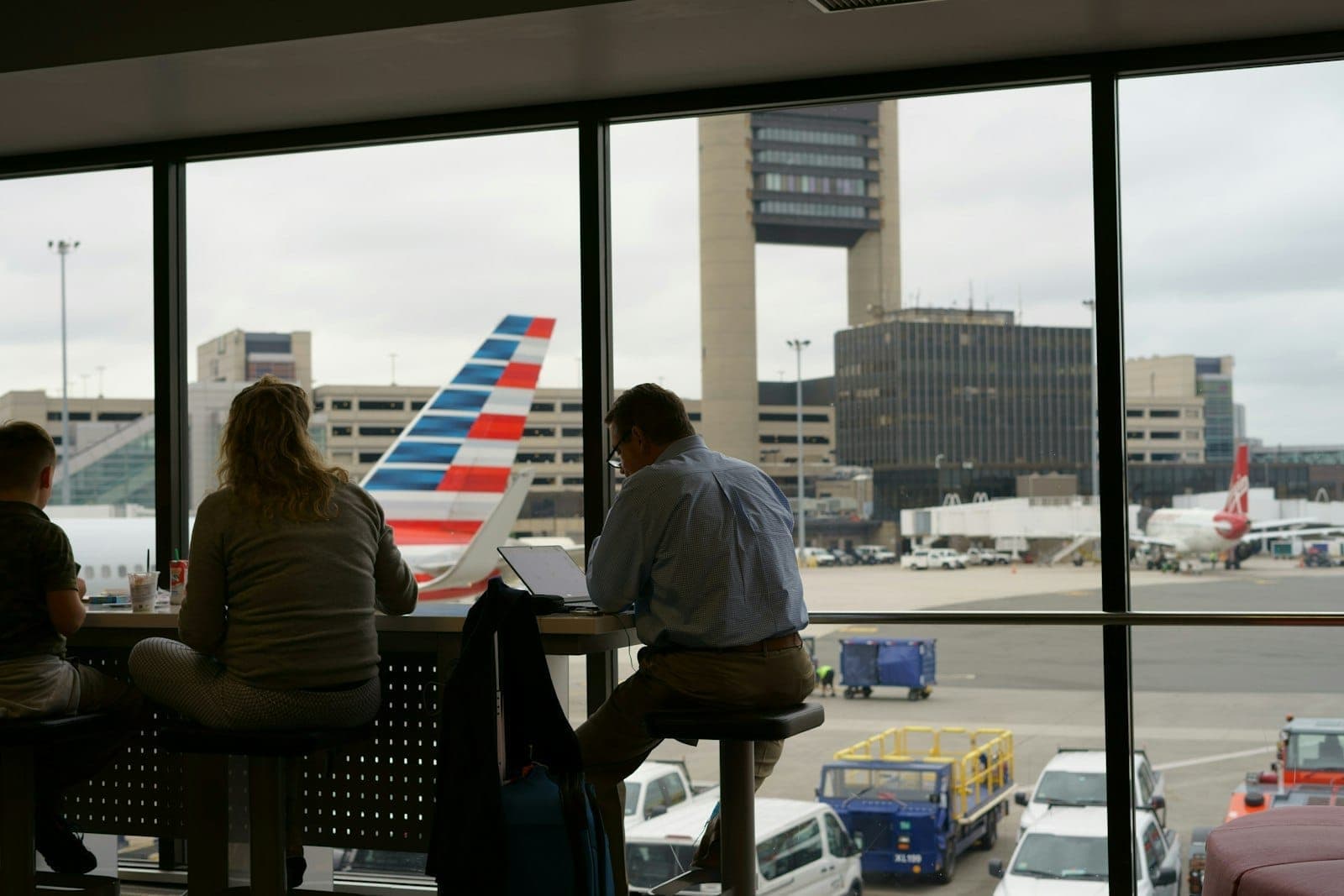 Des passagers assis près d'une grande fenêtre donnant sur le tarmac de l'aéroport, un avion d'American Airlines et la tour de contrôle de l'aéroport Boston Logan.