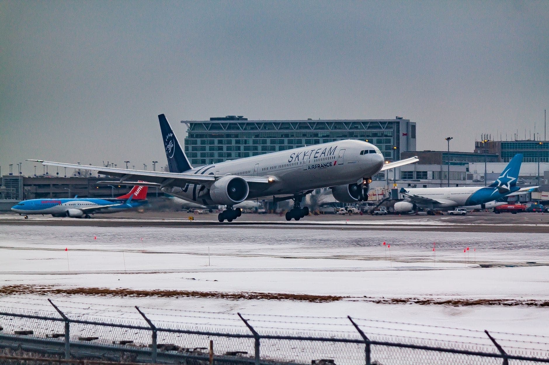 Piste d'aéroport enneigée avec un avion d'Air France atterrissant et des terminaux en arrière-plan, météo hivernale visible.