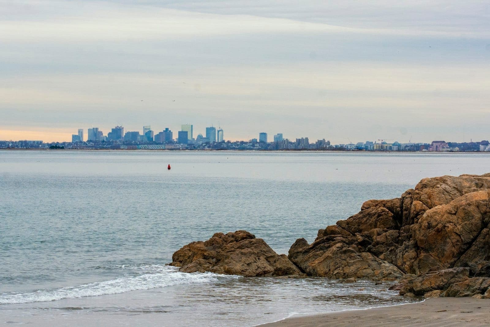 Vue d'une plage rocheuse avec la skyline de Boston au loin sous un ciel nuageux, de douces vagues venant lécher le rivage.