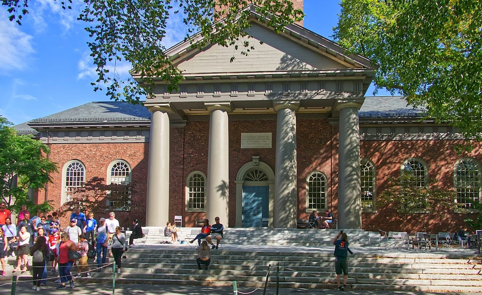 Un bâtiment classique du campus de Harvard avec des colonnes, une façade en brique et des étudiants et visiteurs rassemblés sur les marches au soleil.