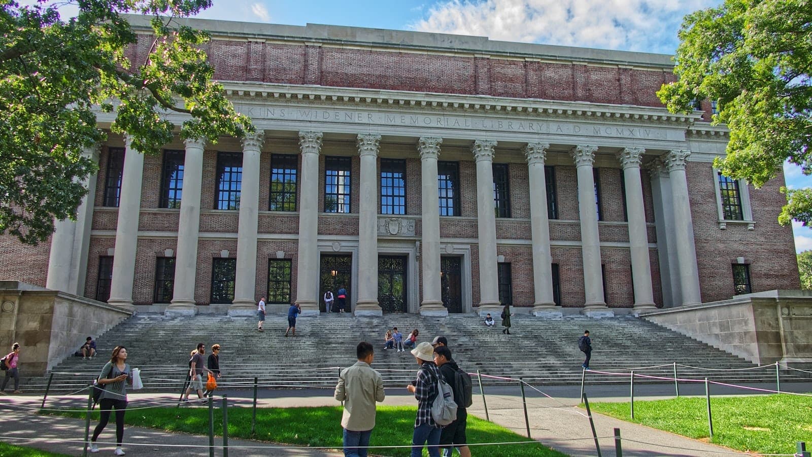 Vue de face de l'emblématique bibliothèque de Harvard avec ses grandes colonnes et des visiteurs sur les marches, encadrée d'arbres verdoyants en pleine lumière.