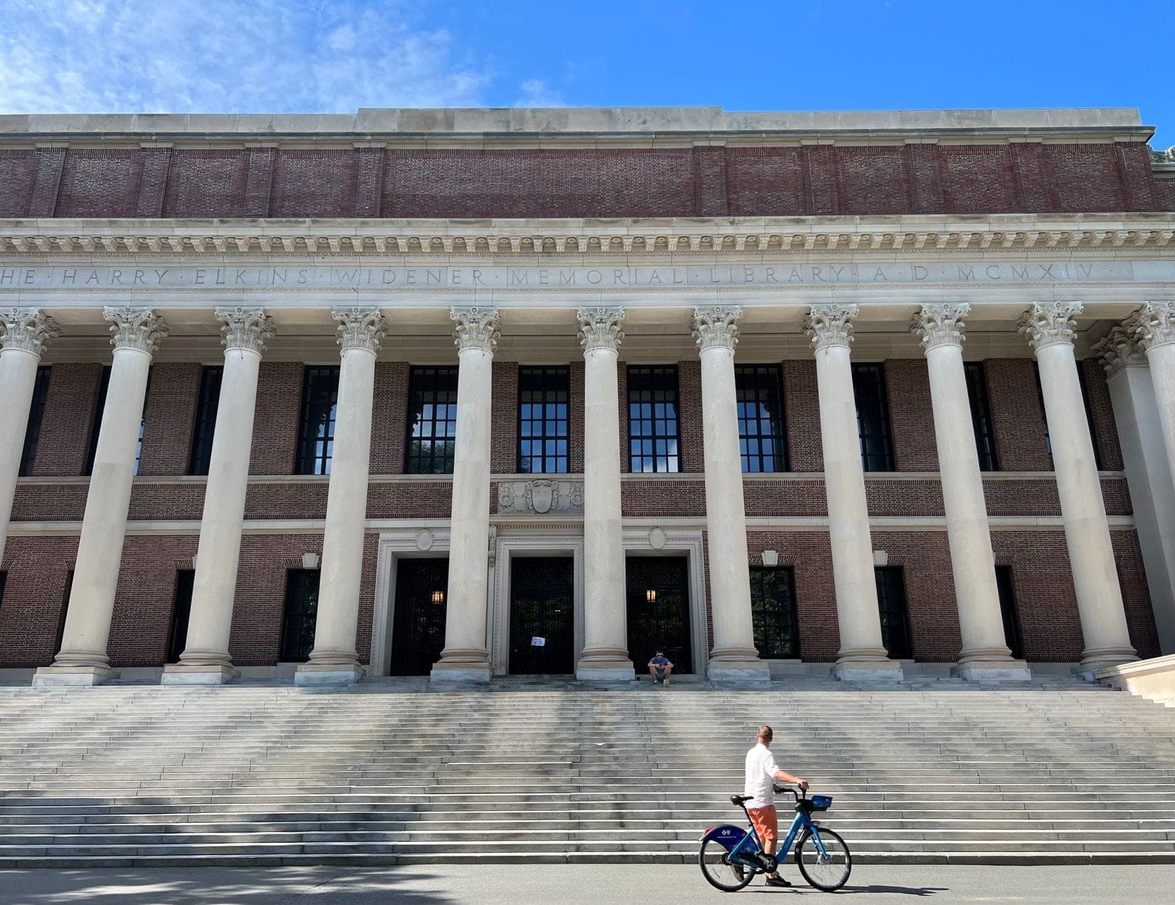 Une personne fait du vélo devant la Widener Library de Harvard, avec ses colonnes emblématiques et ses larges marches sous un ciel bleu dégagé.