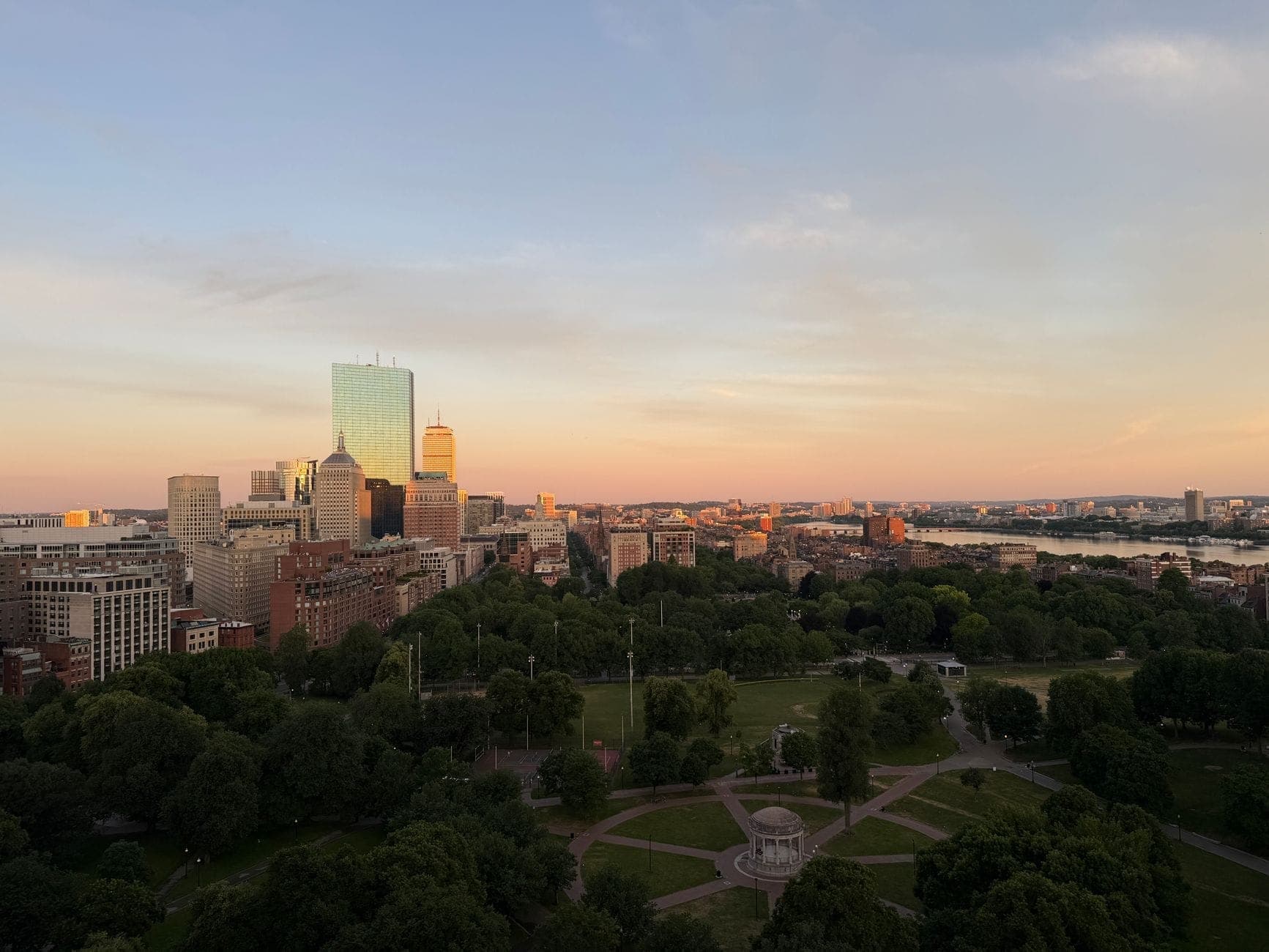 Vue aérienne de Boston Common au coucher du soleil, avec ses vastes espaces verts, ses allées historiques, la skyline du centre-ville et le Charles River au loin.