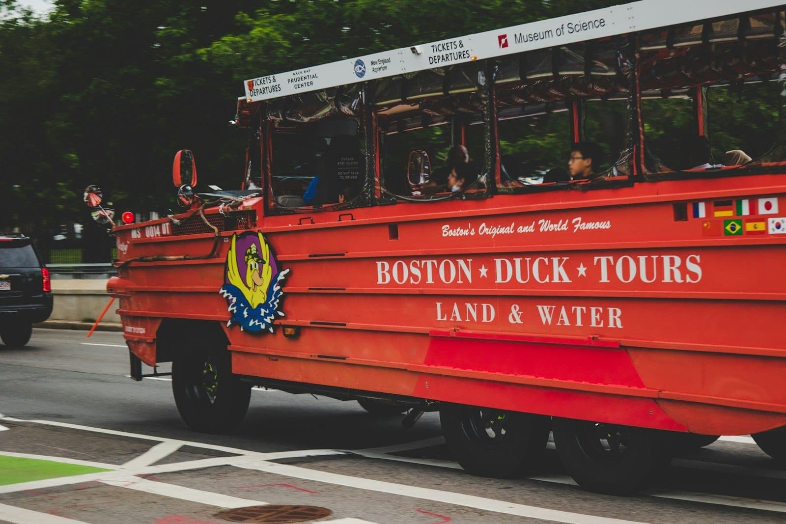 Véhicule amphibie rouge vif de Boston Duck Tours roulant dans une rue de la ville avec des passagers et une signalisation de visite visibles, des arbres en arrière-plan.