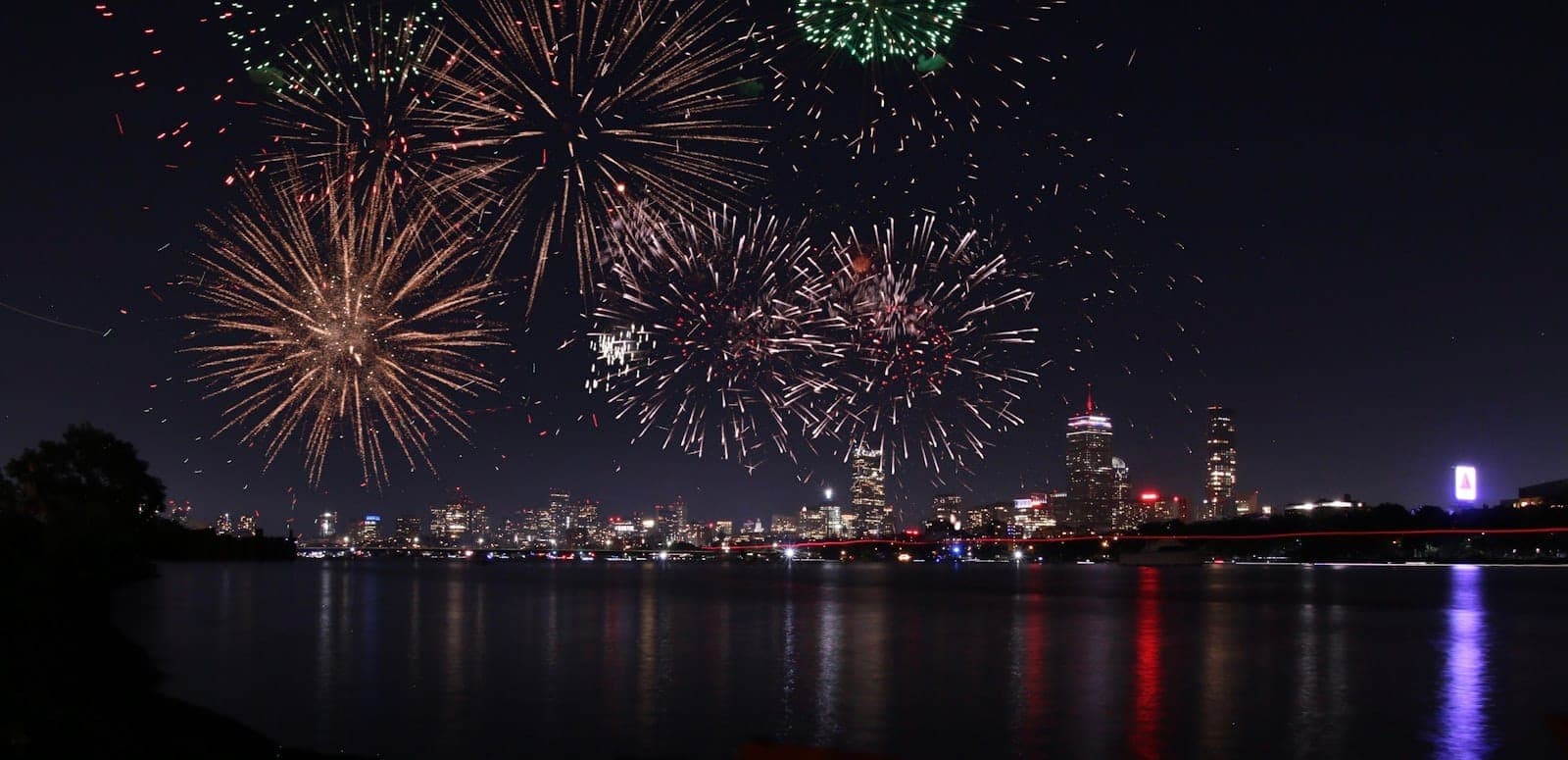 De grands feux d'artifice colorés illuminent le ciel nocturne au-dessus des toits de Boston et du Charles River, avec les lumières de la ville se reflétant sur l'eau.