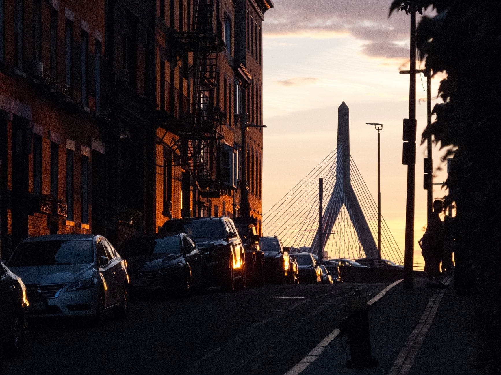 Rue d'un quartier de Boston au coucher du soleil avec des voitures garées, des bâtiments historiques en brique et une vue sur le Zakim Bridge en arrière-plan.