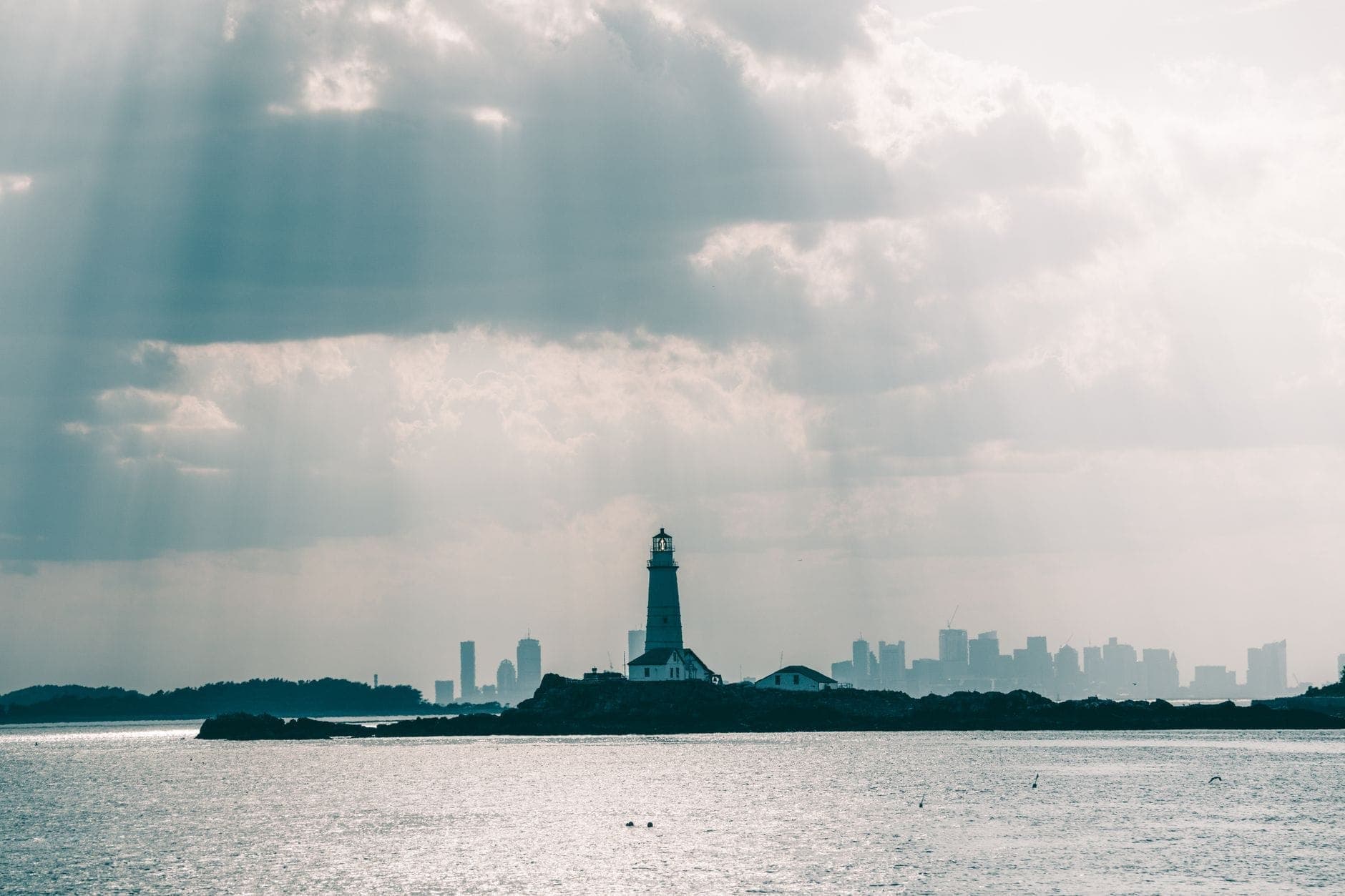 Phare sur une île dans Boston Harbor avec la skyline de la ville visible en arrière-plan et des rayons de soleil traversant les nuages.