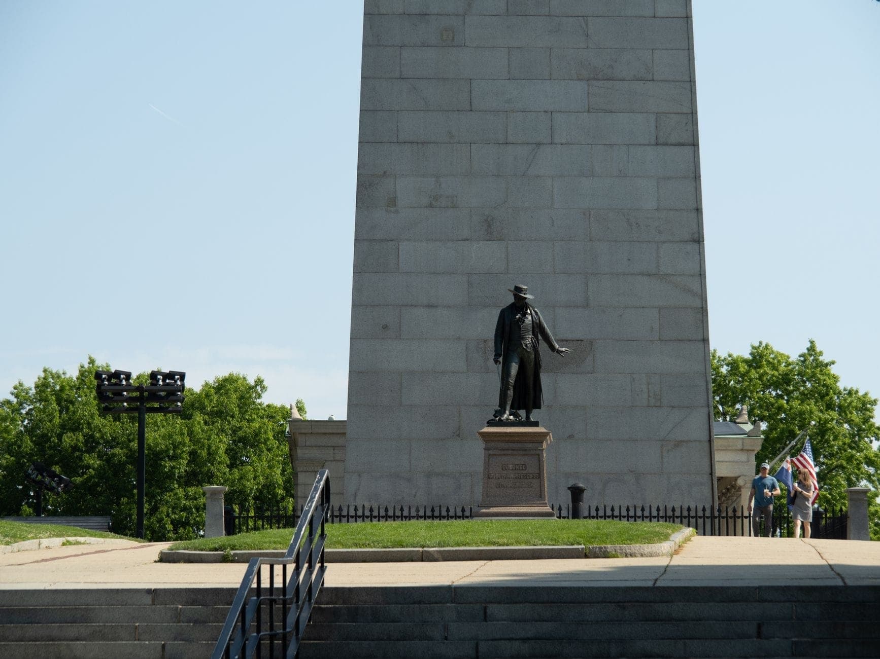 Le Bunker Hill Monument avec une statue et quelques visiteurs tenant des drapeaux américains à sa base par une journée ensoleillée.
