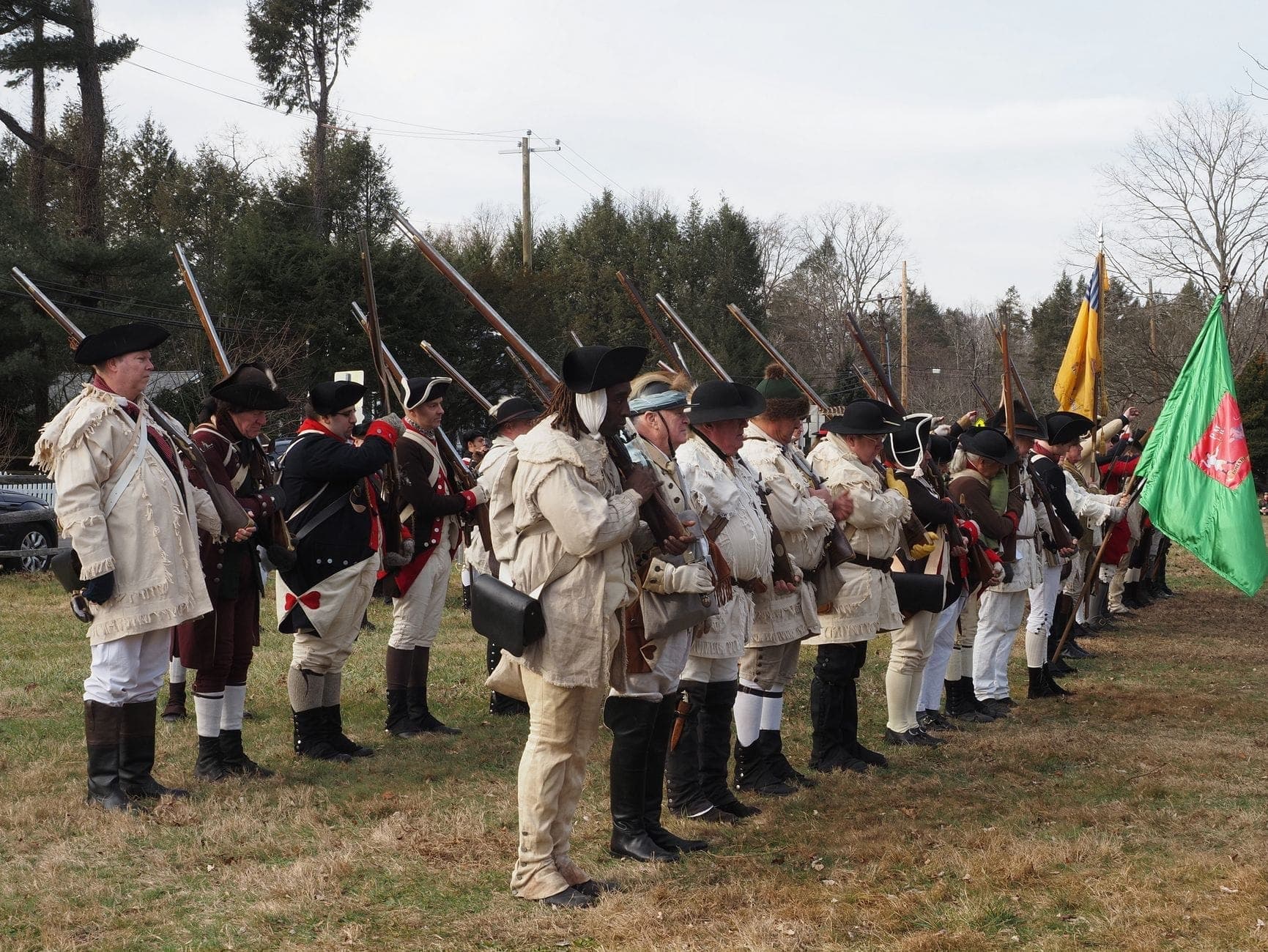 Groupe de reconstituteurs de la Guerre Révolutionnaire en tenue coloniale debout avec des mousquets sur une zone herbeuse, évoquant le champ de bataille de Lexington ou Concord.