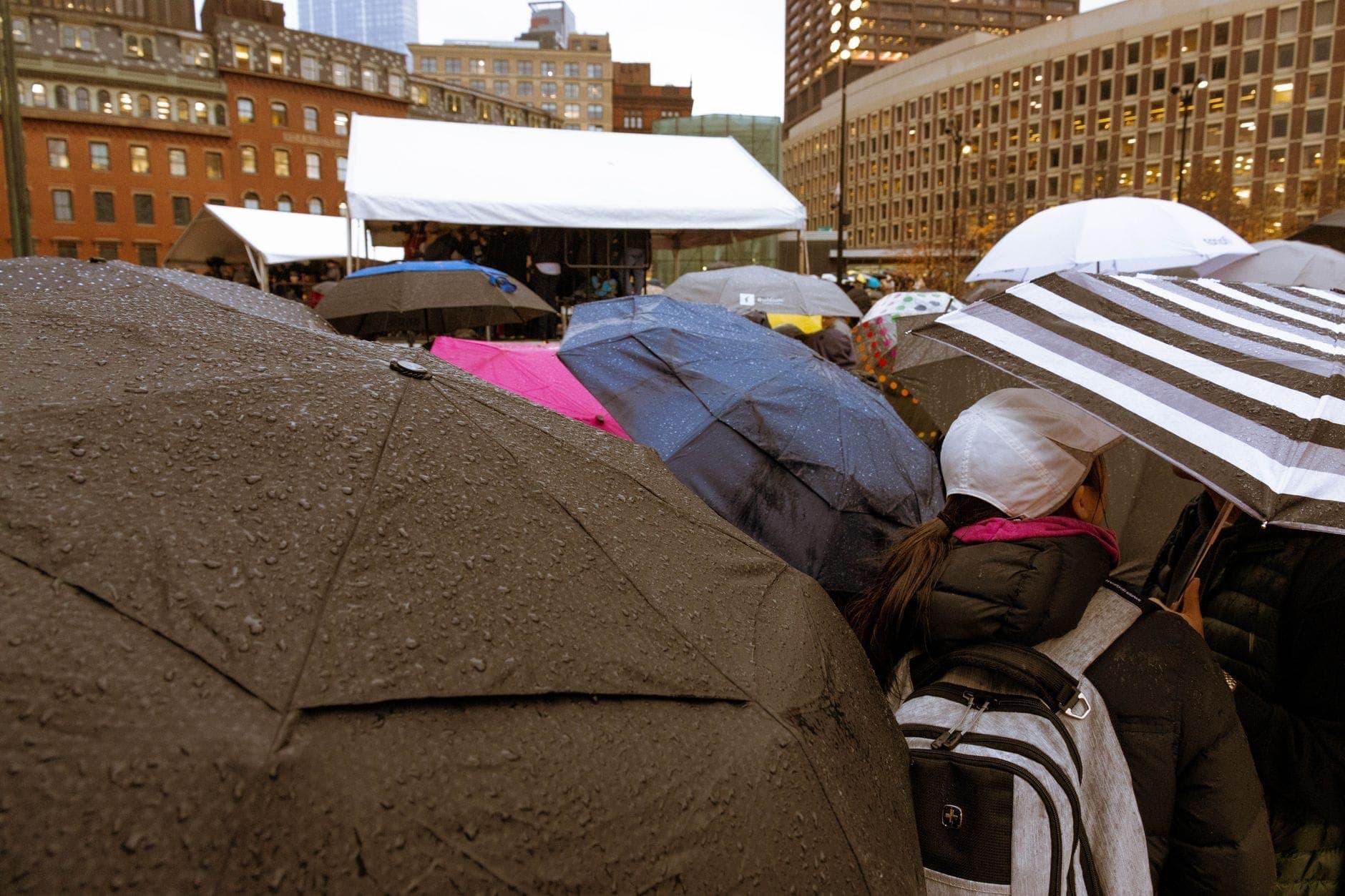Foule avec des parapluies lors d'un événement en plein air sur une place de Boston avec des bâtiments reconnaissables du centre-ville en arrière-plan.