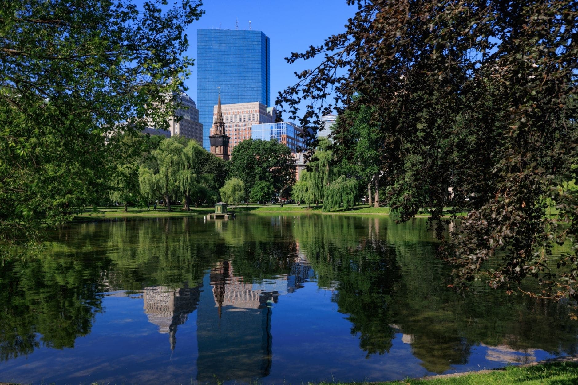 Boston Public Garden en été avec un étang réfléchissant, des arbres verdoyants et la skyline de la ville en arrière-plan sous un ciel bleu dégagé.