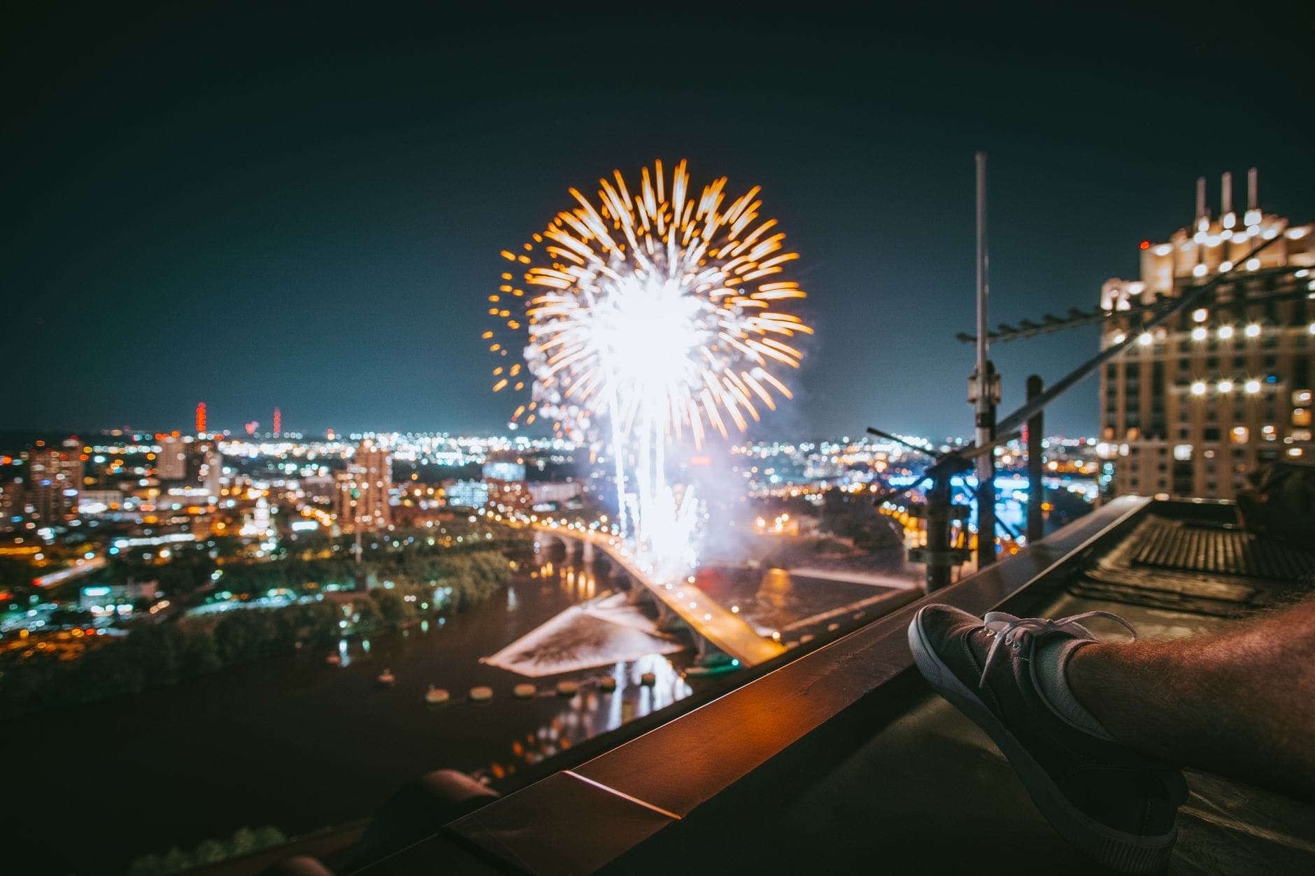 Vue nocturne de feux d'artifice éclatant au-dessus de la rivière avec un paysage urbain illuminé et un pont, vus depuis un toit de Boston.