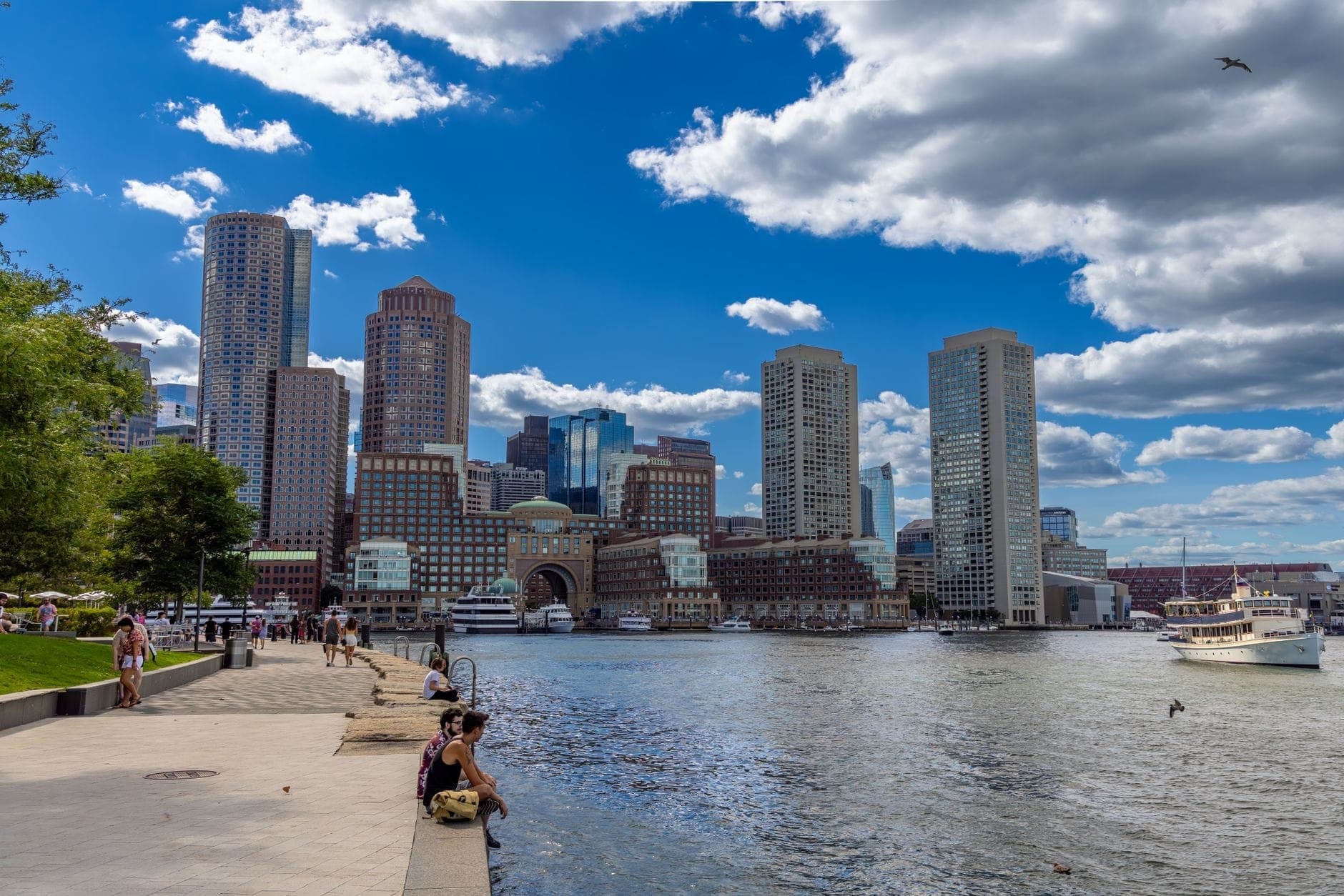 Des personnes profitant du front de mer de Boston par une belle journée ensoleillée, avec ciel bleu, skyline de la ville, port et bateaux en arrière-plan.