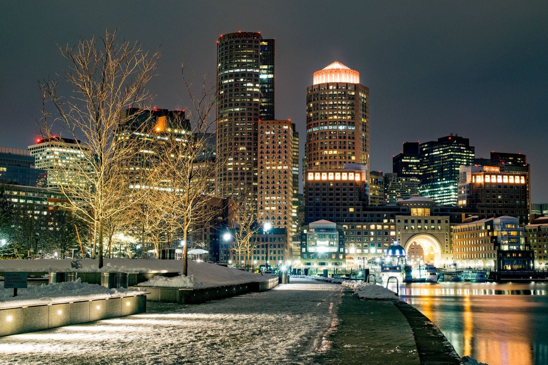 Le centre-ville de Boston illuminé la nuit avec de la neige au sol, les lumières de la ville se reflétant sur l'eau dans une atmosphère hivernale saisissante.
