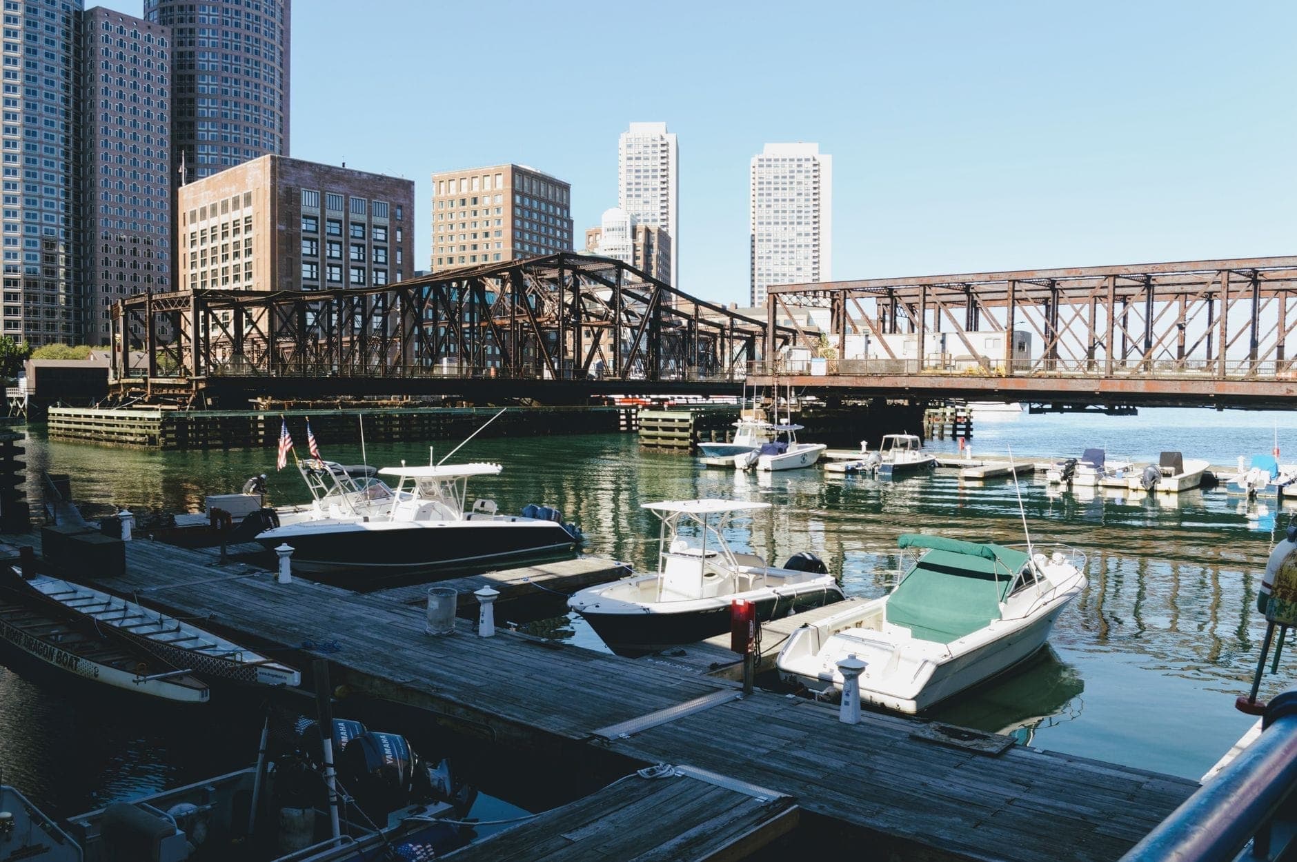 Vue du front de mer de Boston avec des bateaux de luxe amarrés dans une marina, des immeubles formant la skyline de la ville et un pont historique en arrière-plan.