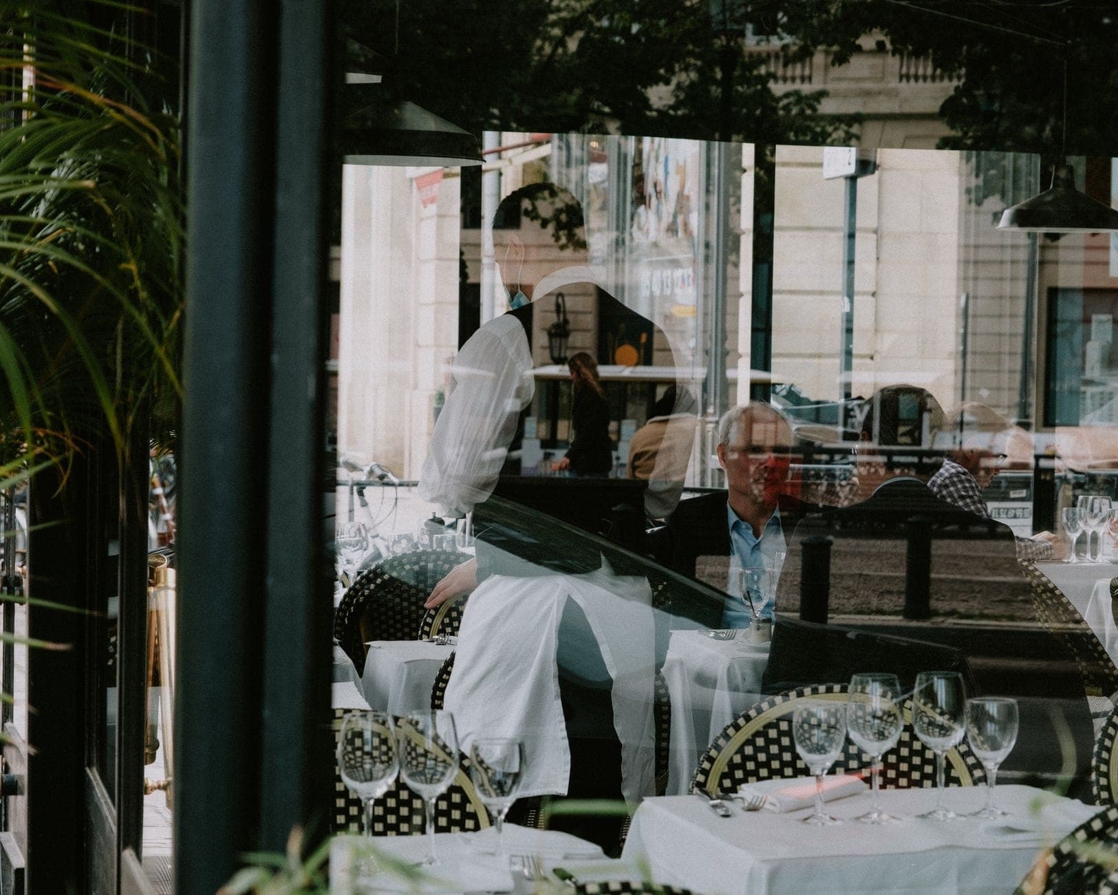 Intérieur élégant d'un restaurant avec des nappes blanches, des verres à vin et un serveur s'occupant des clients assis près de grandes baies vitrées.