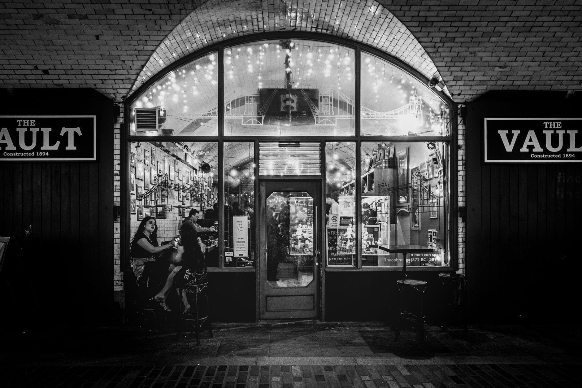 Photo en noir et blanc de l'entrée du bar The Vault à Boston la nuit, avec des personnes assises dehors sous des guirlandes lumineuses.