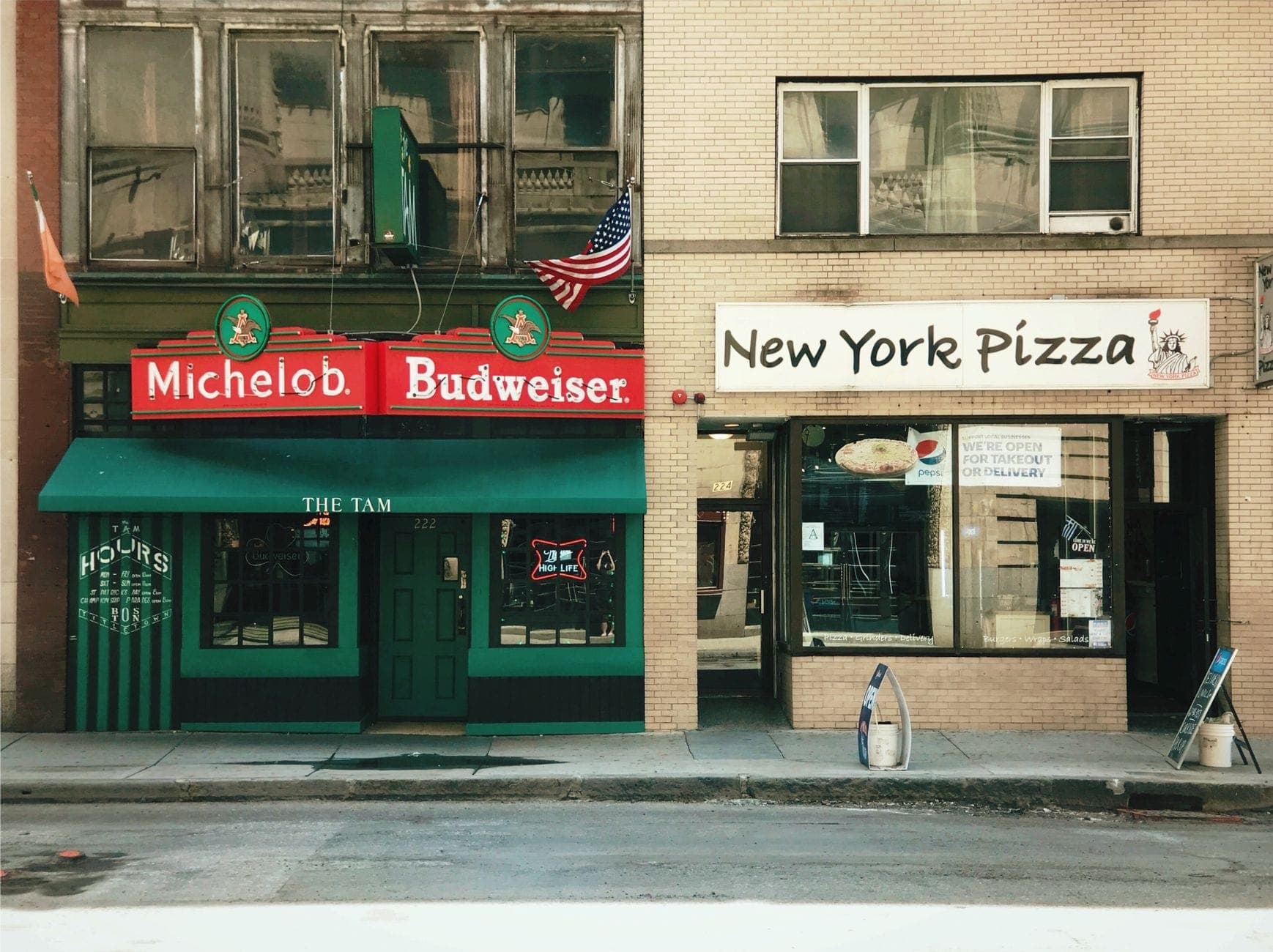 Devantures du bar The Tam et de New York Pizza dans une rue de la ville, avec des enseignes vertes et rouges et une atmosphère classique de quartier.